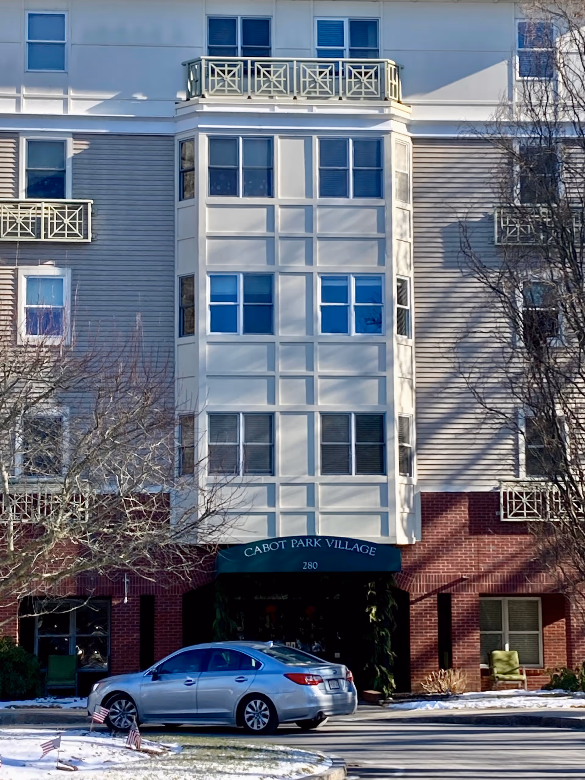 Front exterior view of a multi-story senior living facility named Cabot Park Village with a silver car parked in front. The building has a combination of brick and light-colored siding with multiple windows and decorative balconies. There is some snow on the ground and leafless trees near the entrance.