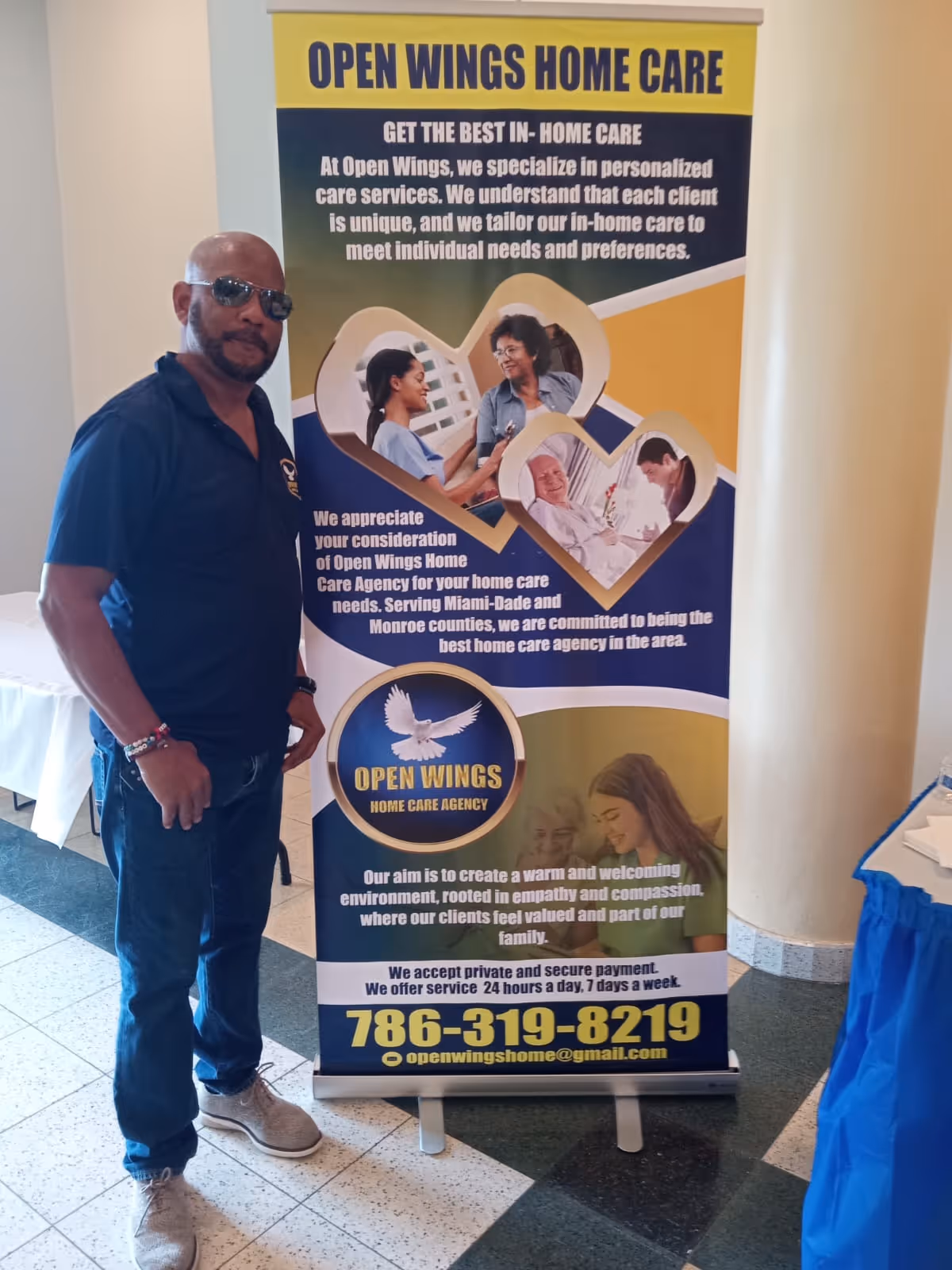 Man stands beside Open Wings Home Care banner indoors.