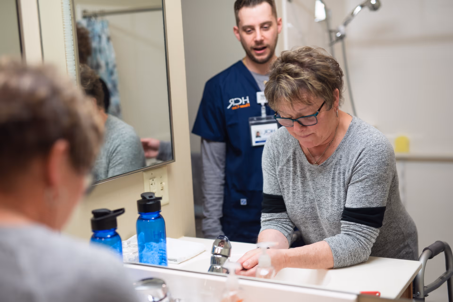 Caregiver supervises a resident washing hands at a bathroom sink in a care facility.