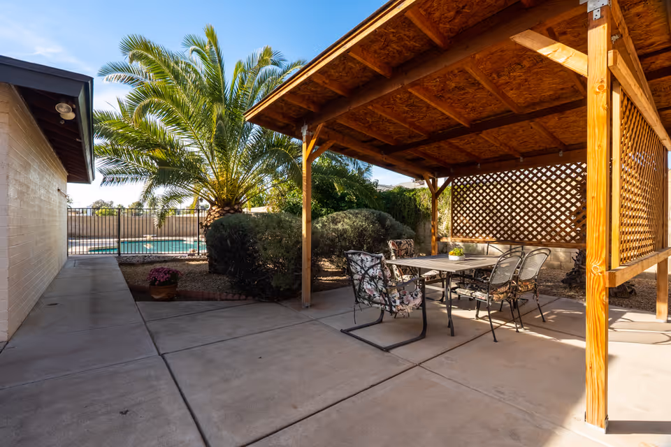 Outdoor patio area with a wooden pergola covering a table and four chairs with floral cushions. There is a palm tree and bushes nearby, and a swimming pool is visible in the background behind a black metal fence.