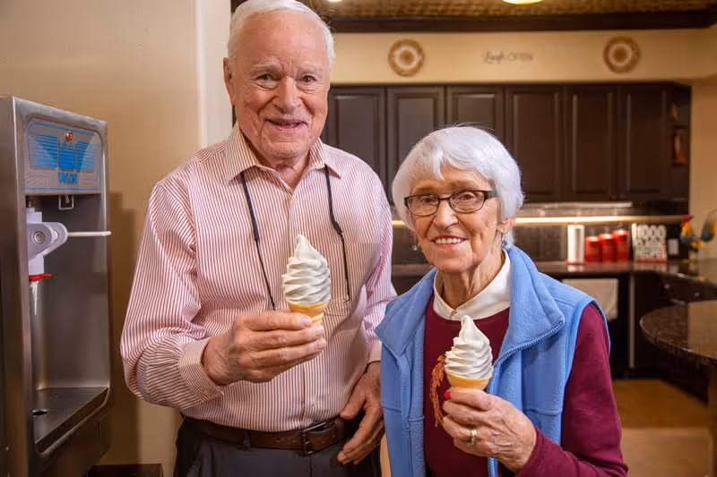 An elderly man and woman standing indoors in a kitchen area, each holding a soft serve ice cream cone and smiling at the camera. The kitchen has dark cabinets and a soft serve ice cream machine is visible on the left side.
