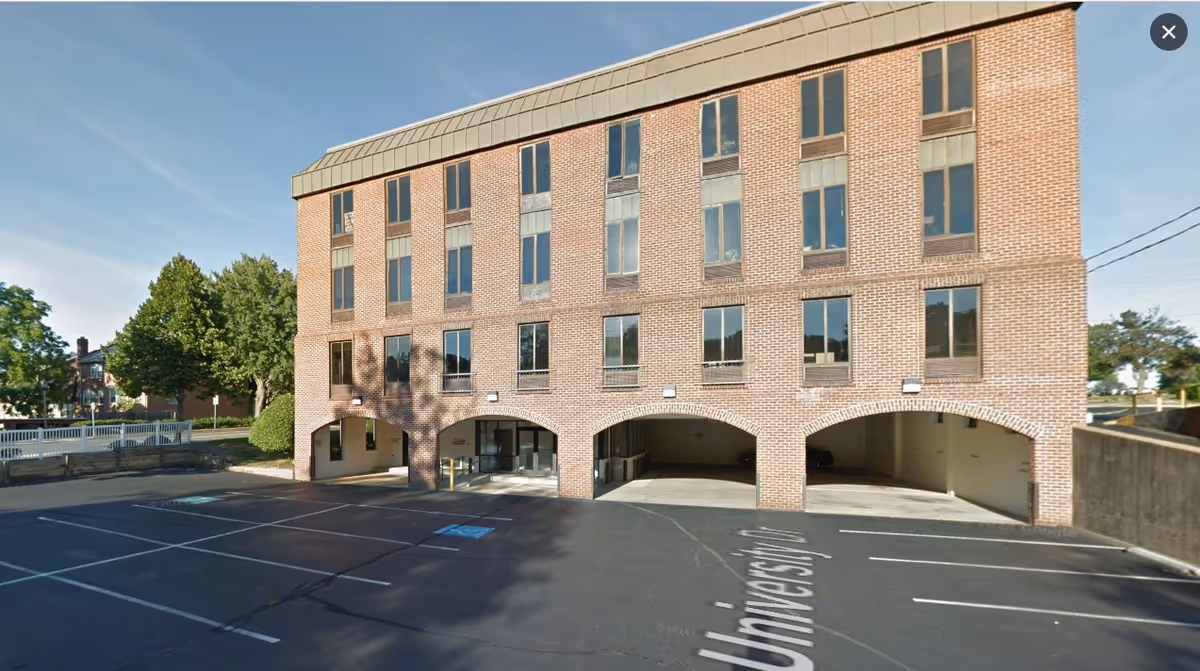 Brick multi-story facility exterior with arched ground-floor openings and a paved parking area in front.