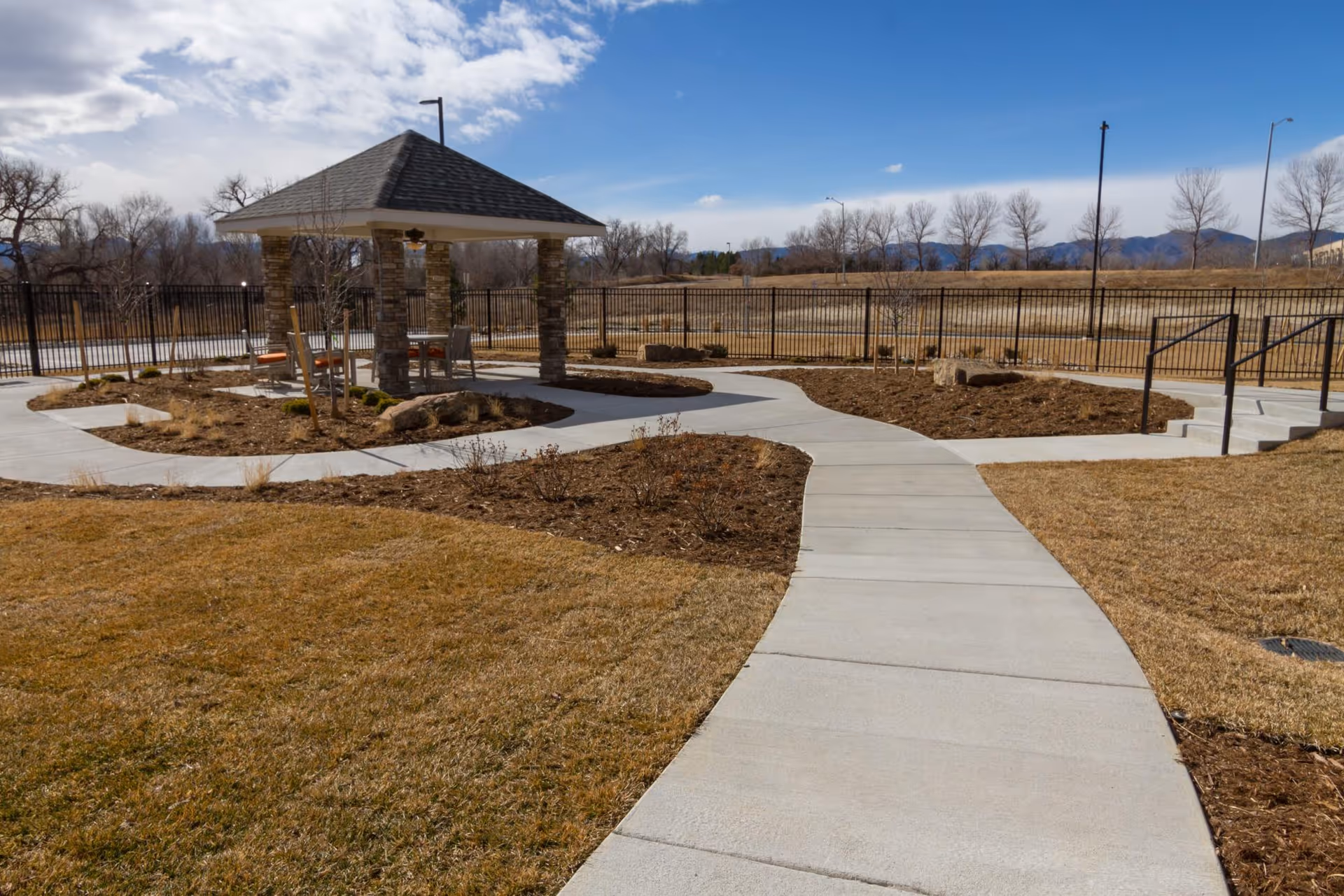 Outdoor area with a concrete pathway leading to a small pavilion with a shingled roof supported by stone pillars. The surrounding landscape includes dry grass, mulched planting beds, and a black metal fence in the background under a partly cloudy sky.