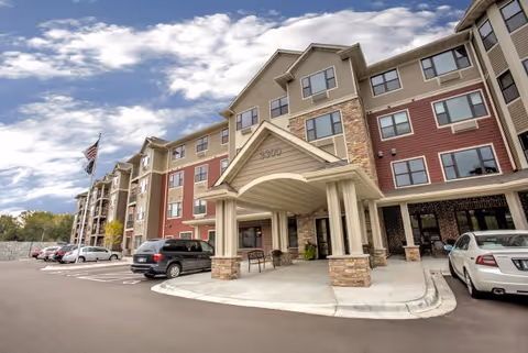 Front exterior view of a multi-story senior living facility building with a covered entrance, several parked cars, and an American flag on a flagpole under a partly cloudy sky.