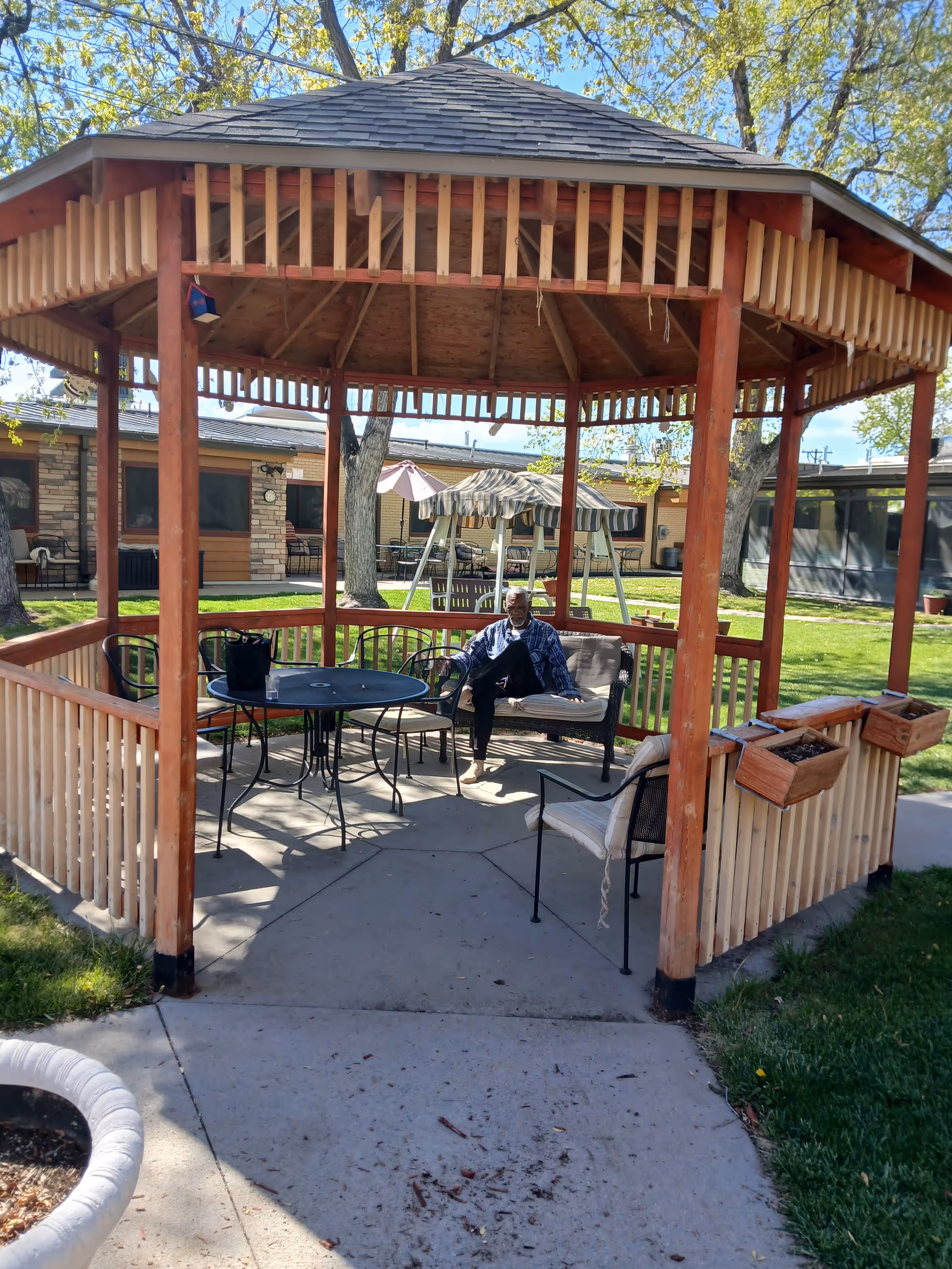 Senior resident relaxing on a couch inside a wooden gazebo in a sunny courtyard.