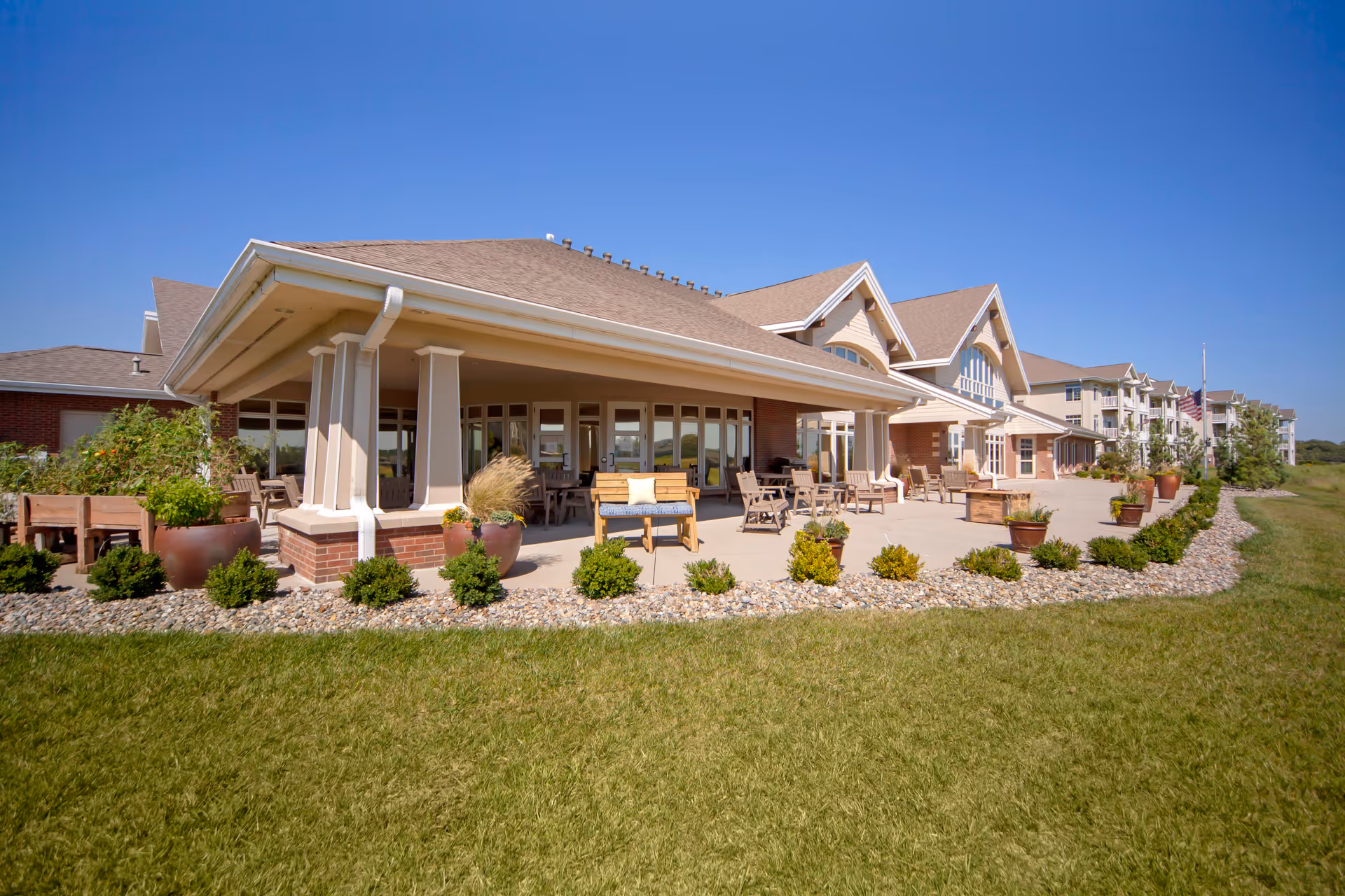 Wide exterior view of an assisted living building with a covered patio, outdoor seating, and landscaped lawn under a clear blue sky.