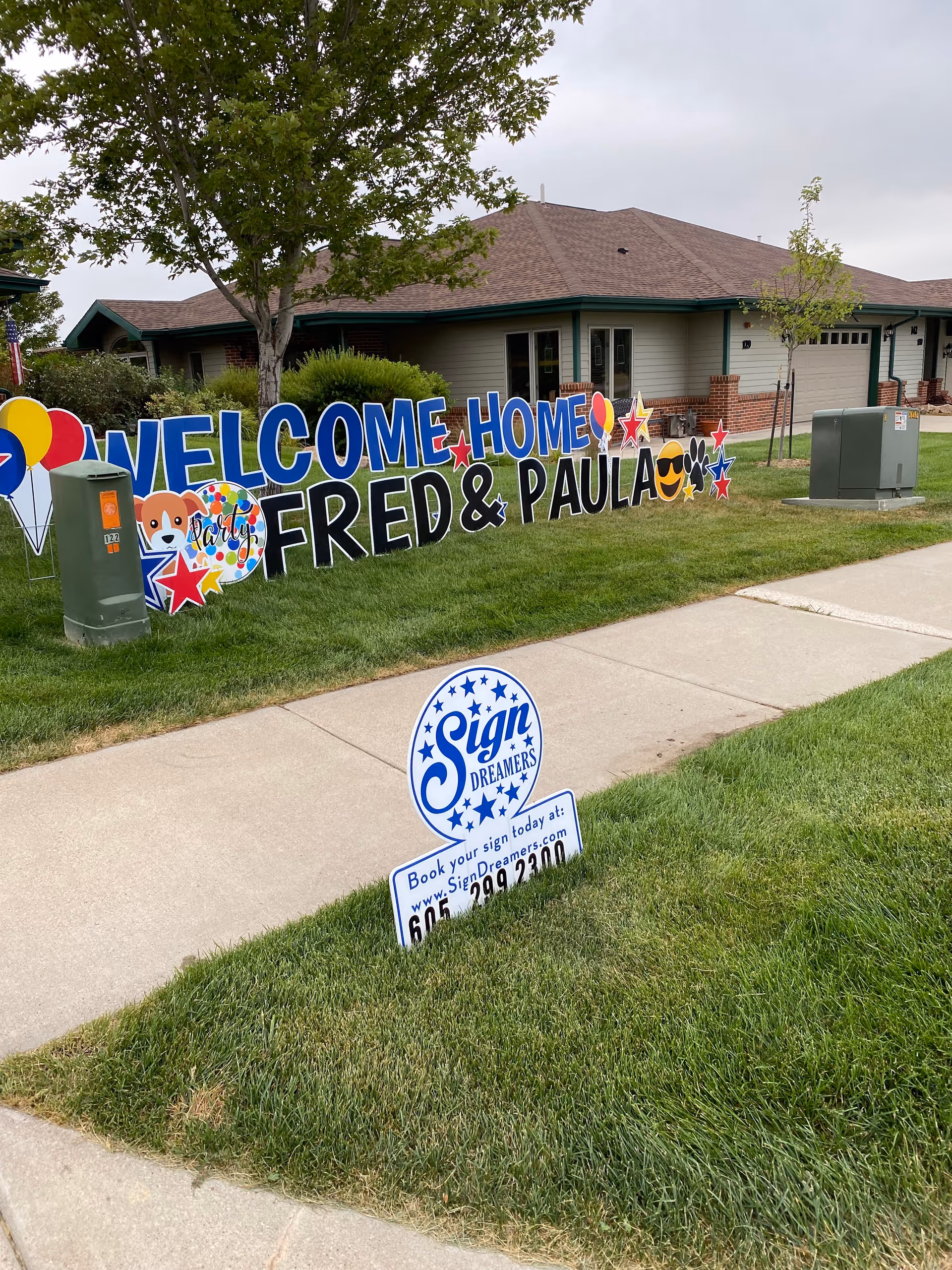 Single-story beige building with large colorful Welcome Home Fred and Paula lawn sign.