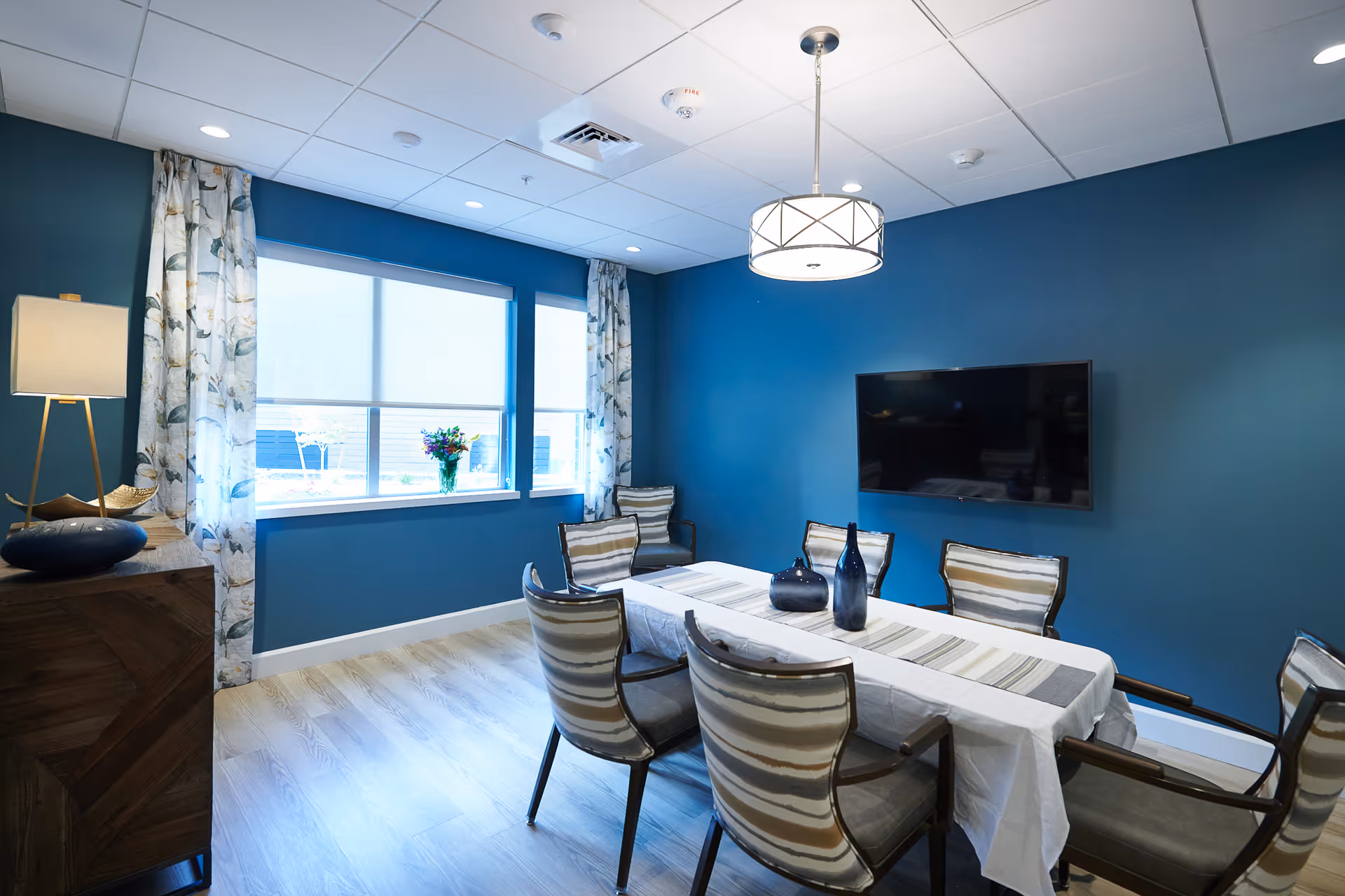 Blue dining room with a long table, striped chairs, window, and wall-mounted TV.