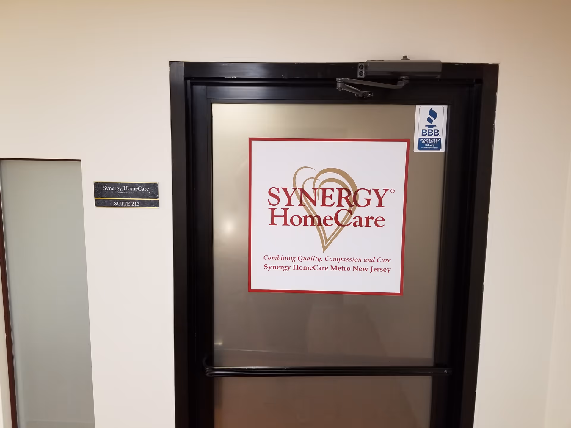 Metal door with Synergy HomeCare sign and suite plaque on beige interior wall.
