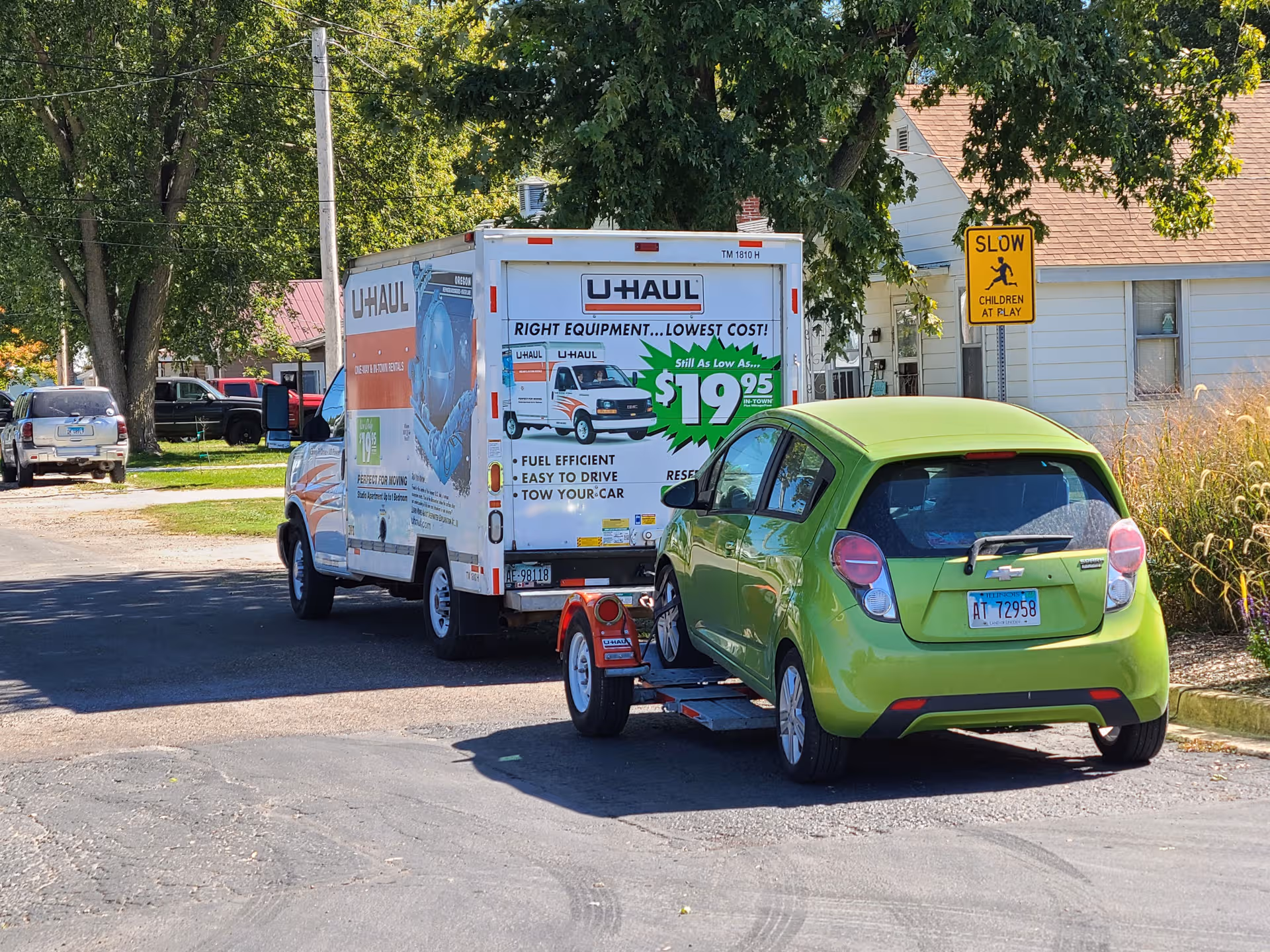A U-Haul truck and a lime-green car on a trailer parked on a residential street.