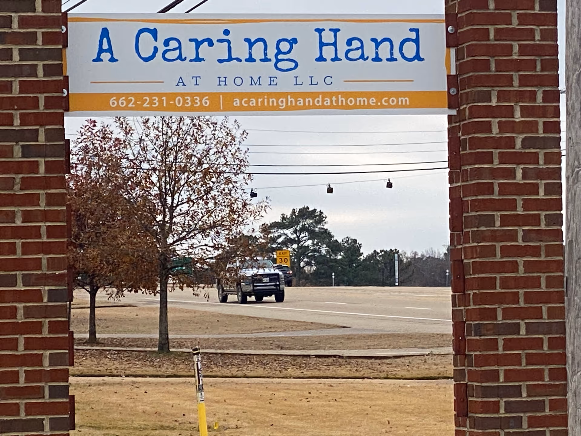 Bricked facility entrance with 'A Caring Hand at Home LLC' sign beside a road.