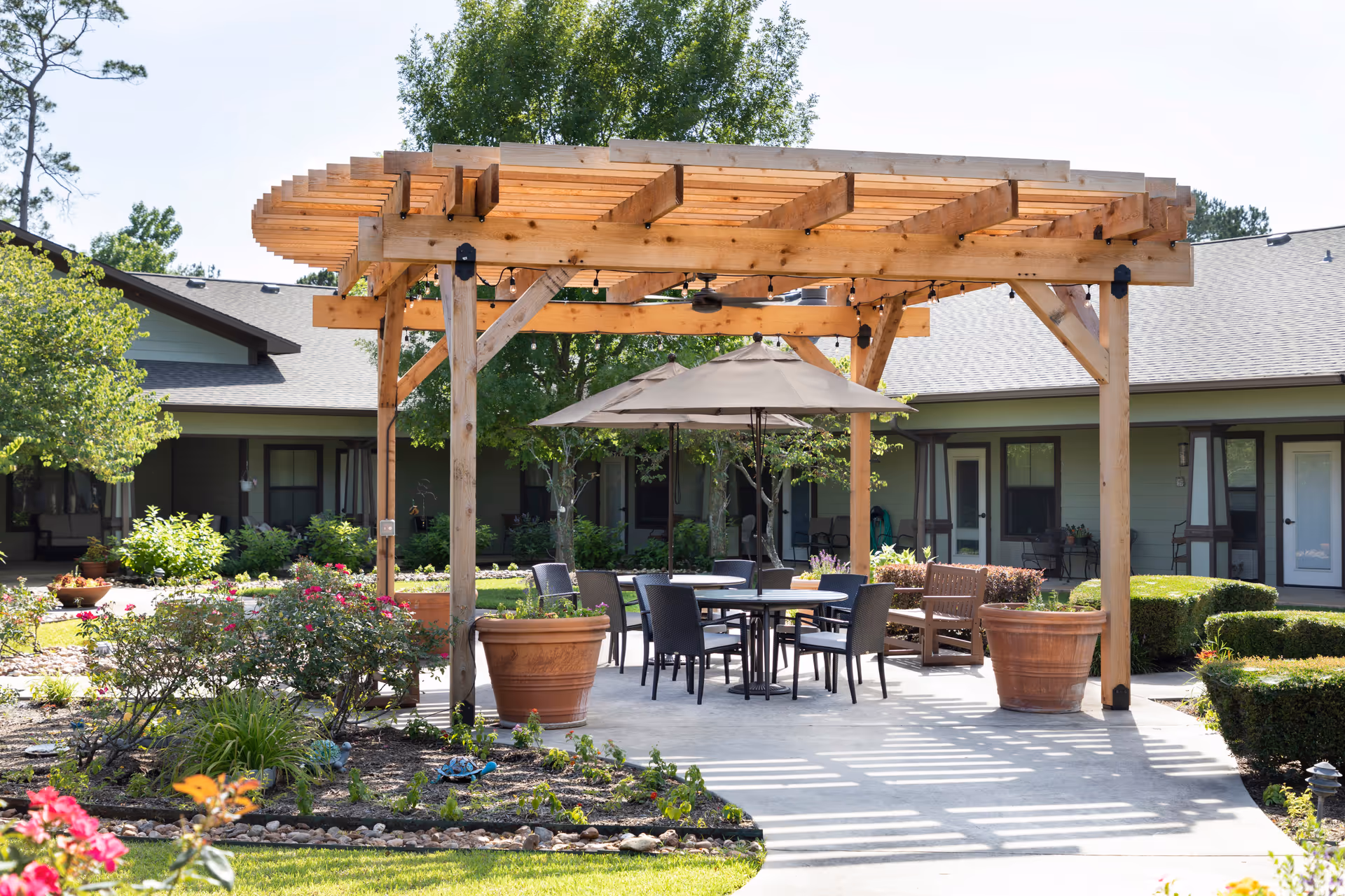 Outdoor seating area at Heritage Oaks Assisted Living and Memory Care featuring a wooden pergola with string lights, a round table with chairs, a large umbrella, potted plants, and surrounding garden beds with flowers and shrubs. The background shows single-story building units with doors and windows.