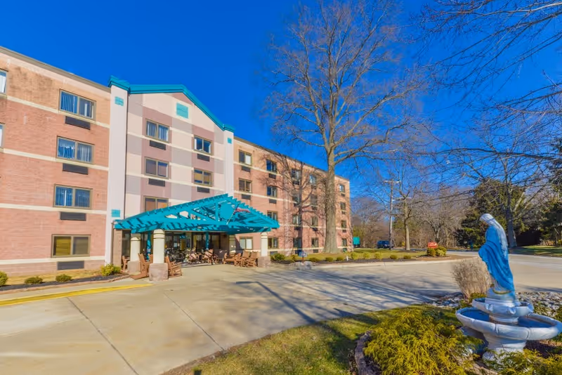 Exterior view of St. Mary Villa for Independent & Retirement Living, showing a multi-story brick building with a teal-colored entrance canopy. There is a paved driveway in front and a statue of a religious figure near a small fountain surrounded by landscaping. Trees without leaves are visible in the background under a clear blue sky.