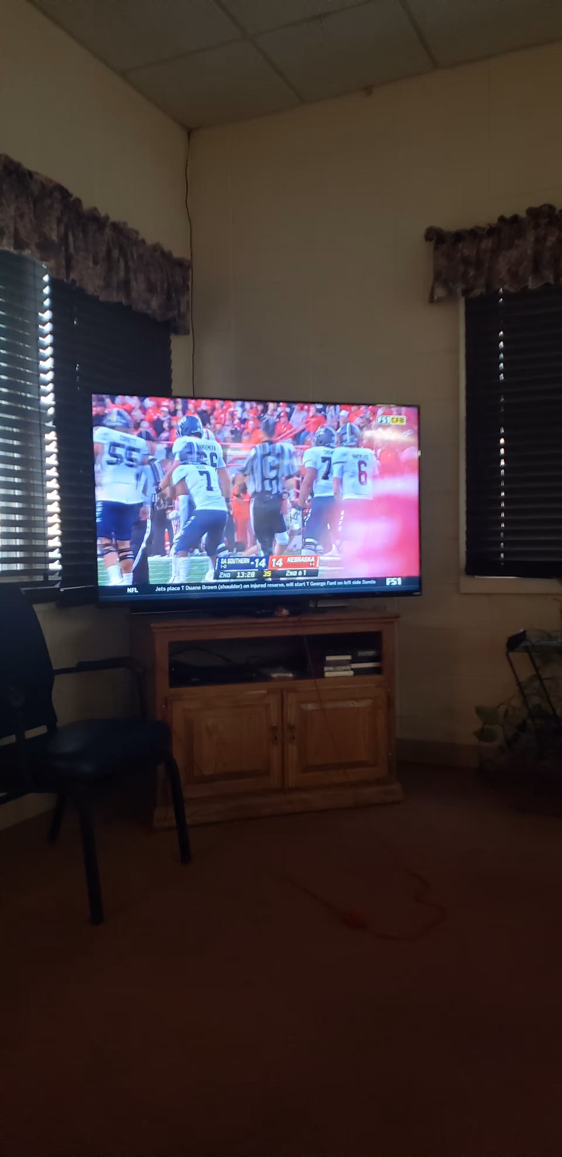TV on a wooden stand in a small living room with two windows and blinds.
