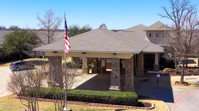 Front exterior view of Keller Oaks Healthcare Center showing a covered entrance with stone pillars, an American flag on a flagpole, surrounding trees, bushes, and a parked car in the driveway.