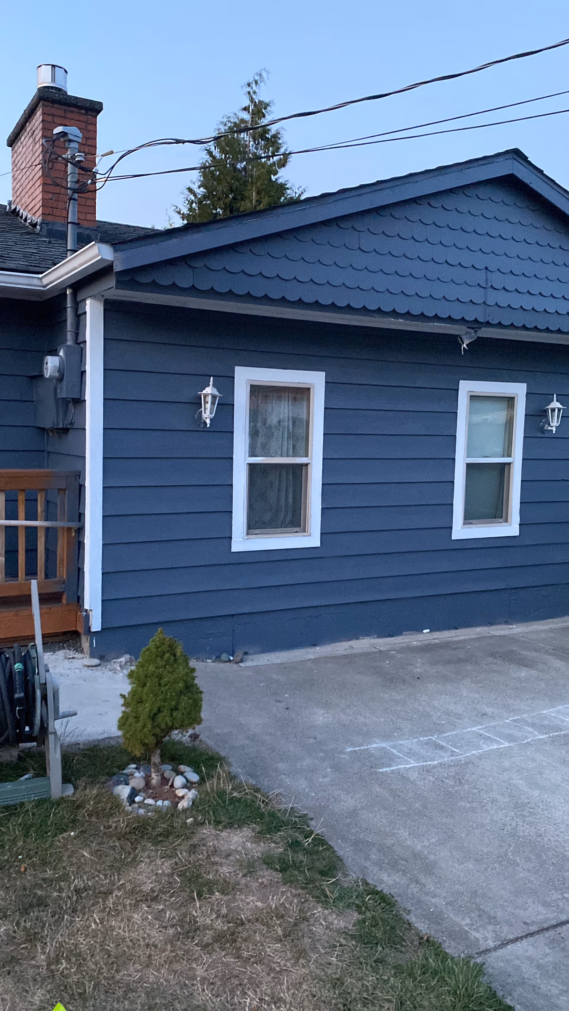 Exterior view of a blue house with white trim, two windows, a small porch with wooden railing, a chimney, and a small landscaped area with a small tree and rocks near a concrete driveway.