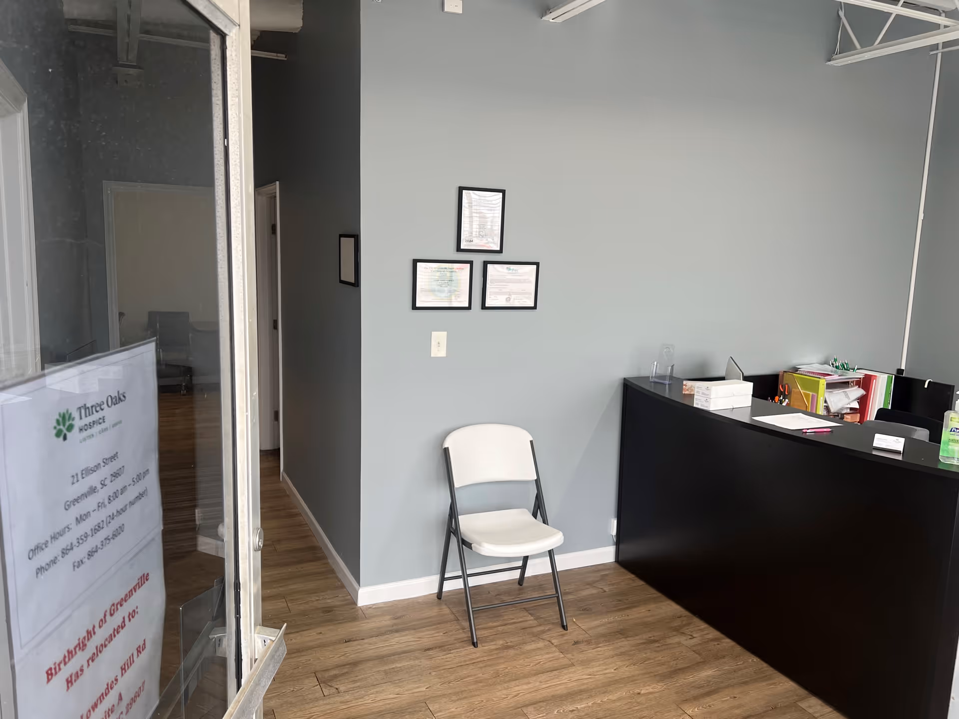 Reception area with a black curved desk, chair, and framed certificates on gray wall.