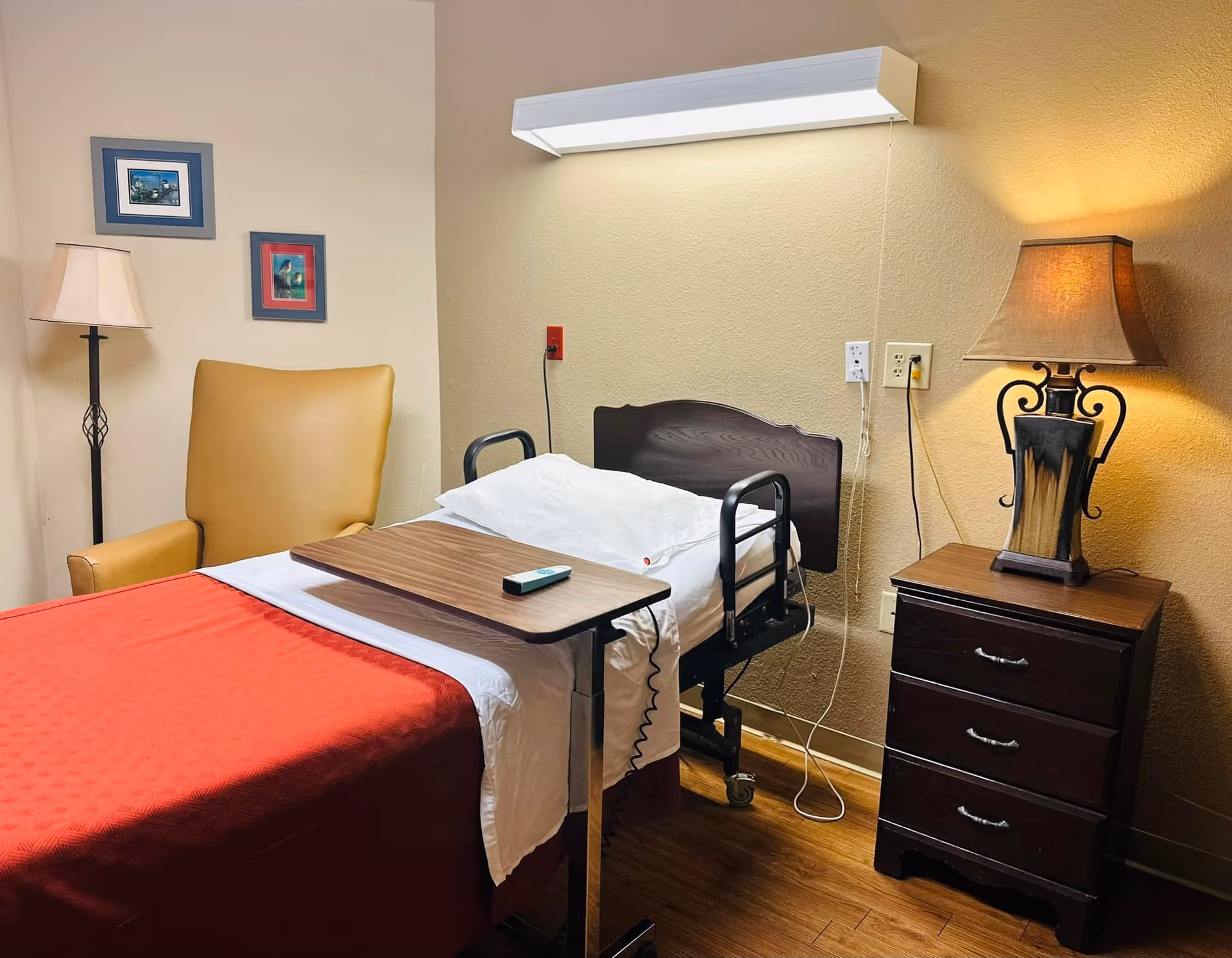 Bedroom in a senior care facility with bed, tray table, chair, dresser, and lamps.