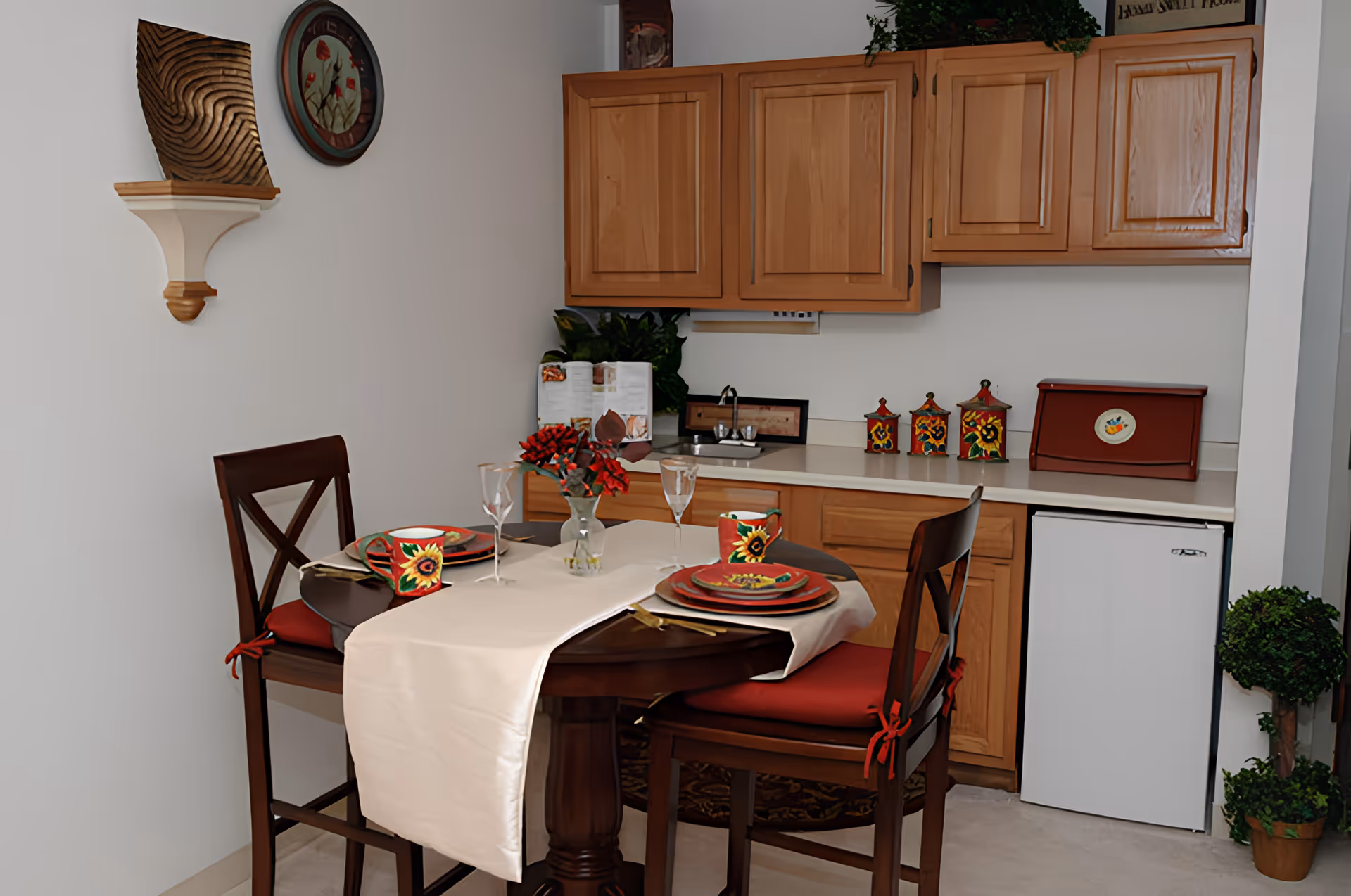 A small dining area with a round table set for two and decorative place settings in front of a kitchenette with wooden cabinets and a mini fridge.