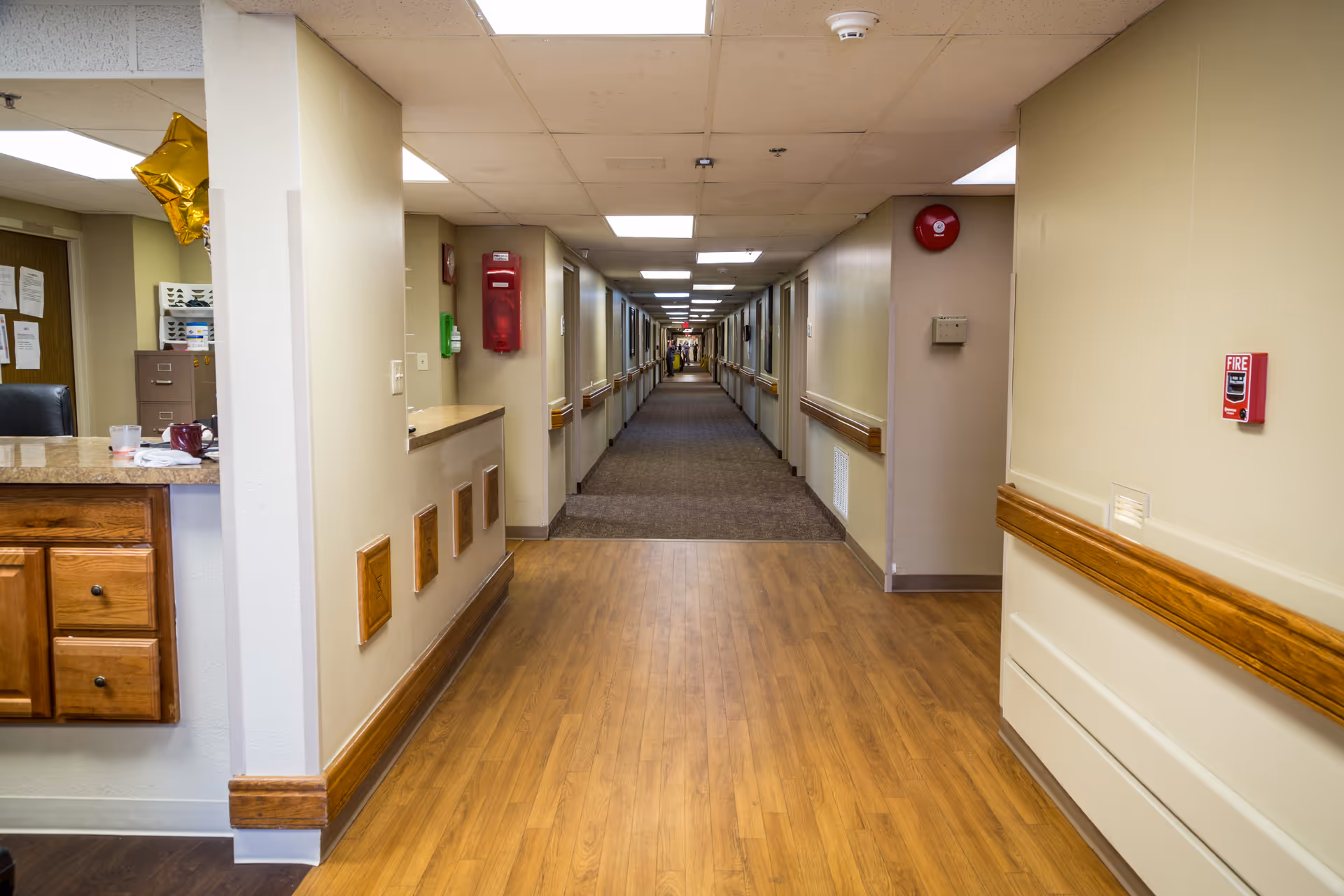 Long hallway inside a senior living facility with wood floor, beige walls, and handrails.