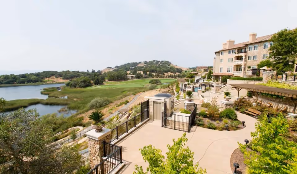 Terraced outdoor patio and landscaped grounds beside a multi-story senior living building overlooking a lake and distant hills.