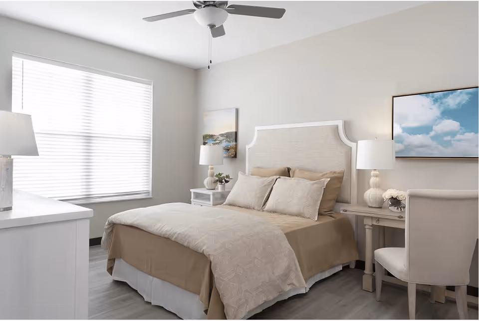 Bright neutral bedroom with a bed, upholstered headboard, nightstands and lamps, a window with blinds, ceiling fan, and a chair at a small desk.
