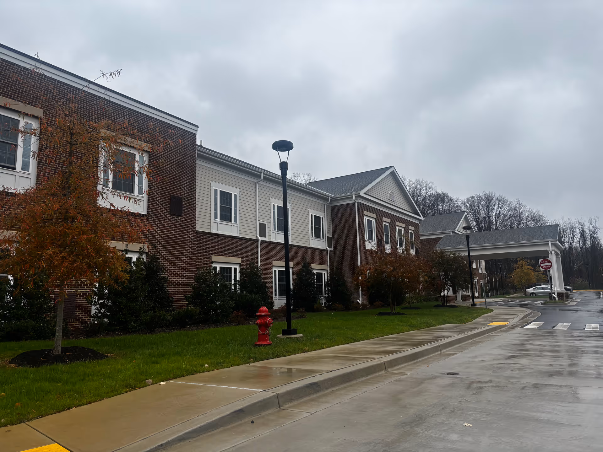 Exterior view of a two-story brick and siding building on a cloudy day with a wet driveway and sidewalk. There are small trees and bushes planted along the building, a red fire hydrant, street lamps, and a covered entrance with a 'Do Not Enter' sign visible.