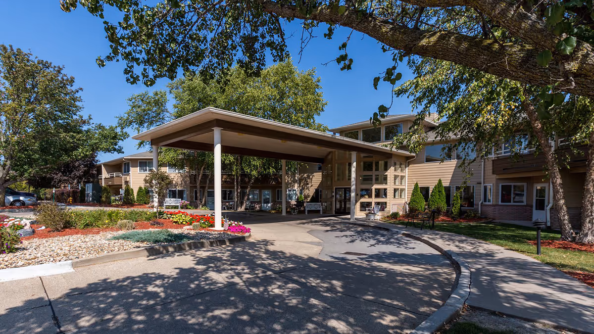 Covered porte-cochère and driveway leading to the front entrance of a two-story residential facility surrounded by trees and landscaping.