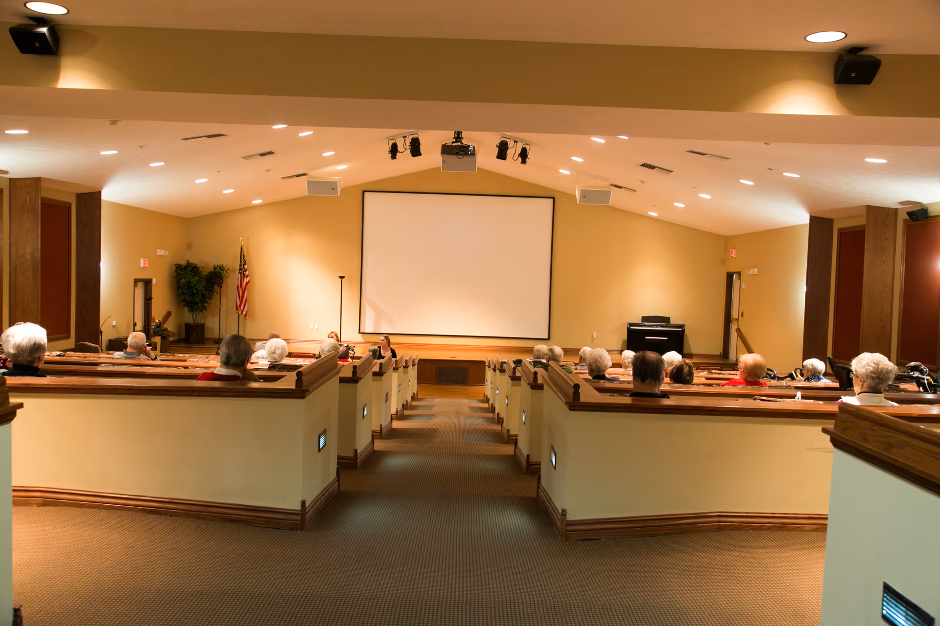 Interior view of a theater-style room with tiered seating occupied by elderly people facing a large blank projection screen. The room has beige walls, carpeted floor, and ceiling lights. An American flag and a piano are visible near the screen.