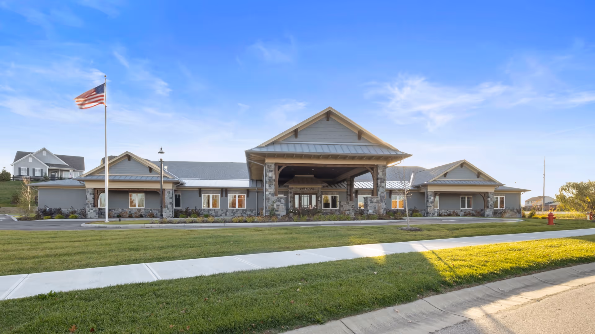 Exterior front view of a single-story senior living facility building with a covered entrance supported by stone pillars, an American flag on a flagpole, well-maintained green lawn, and a clear blue sky.