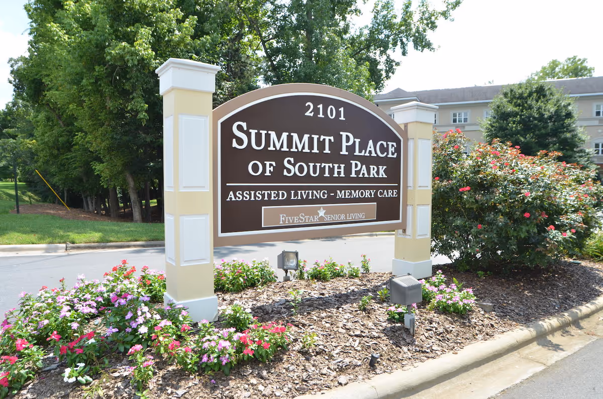 Outdoor view of a large brown and beige sign for Summit Place of South Park, an assisted living and memory care facility, surrounded by flower beds and greenery with a building and trees in the background.