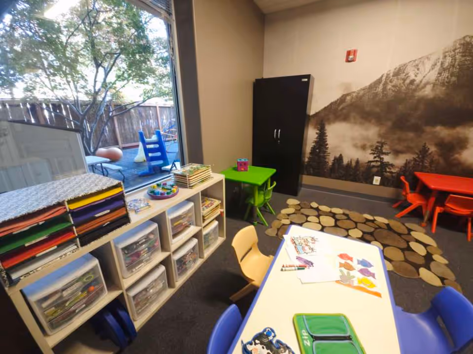 Colorful interior activity room with tables, chairs, and craft shelves near the doorway.