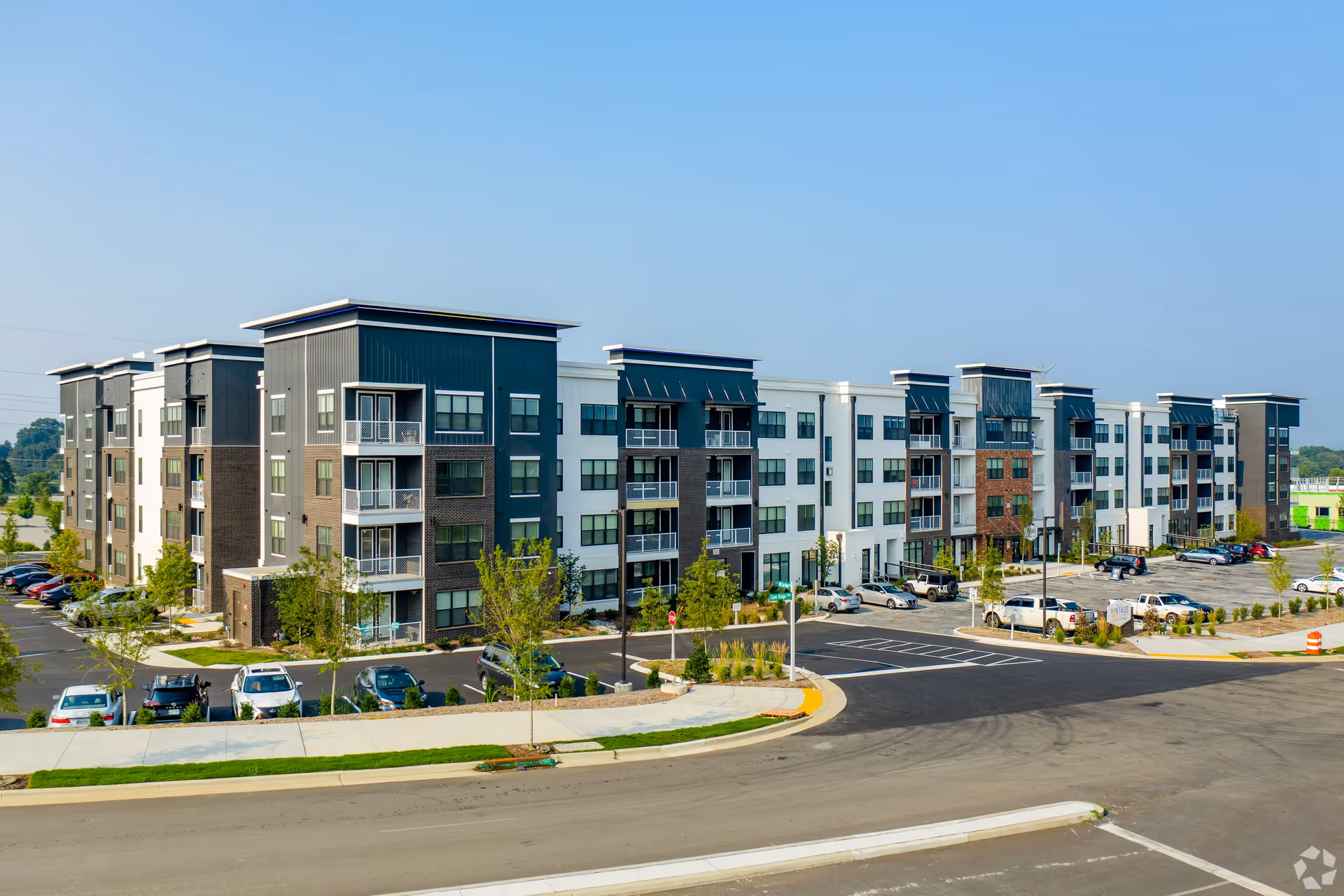 Exterior view of a modern, multi-story apartment building with balconies, surrounded by a parking lot with several parked cars and landscaped greenery under a clear blue sky.