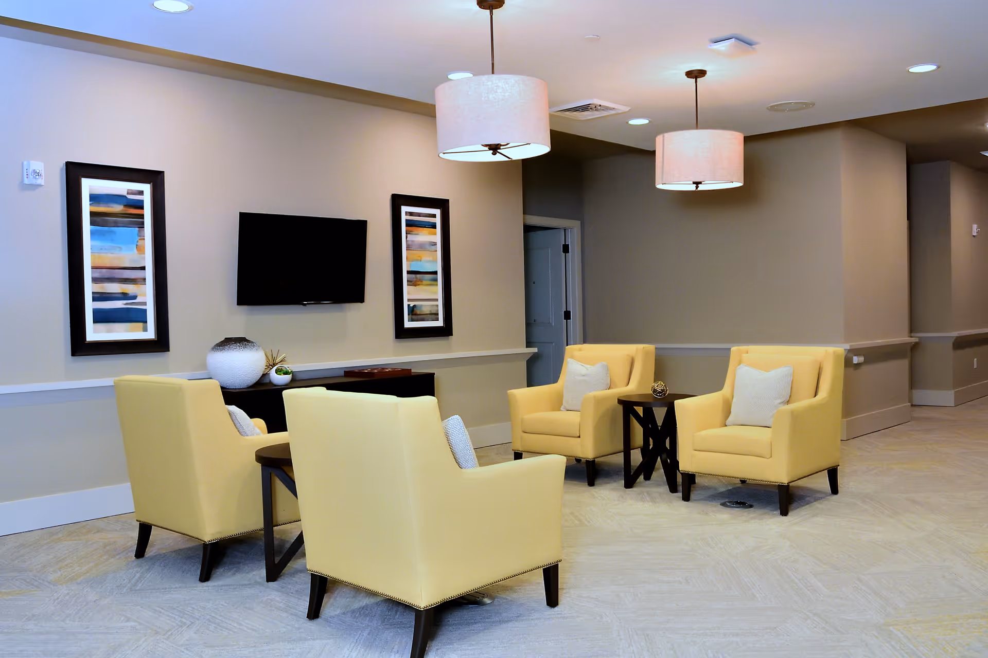 A modern senior living facility common area with four yellow armchairs arranged around two small dark wood tables. The room has beige walls with two abstract framed artworks and a flat-screen TV mounted on the wall. Two pendant lights hang from the ceiling, and the floor is covered with light-colored tiles.