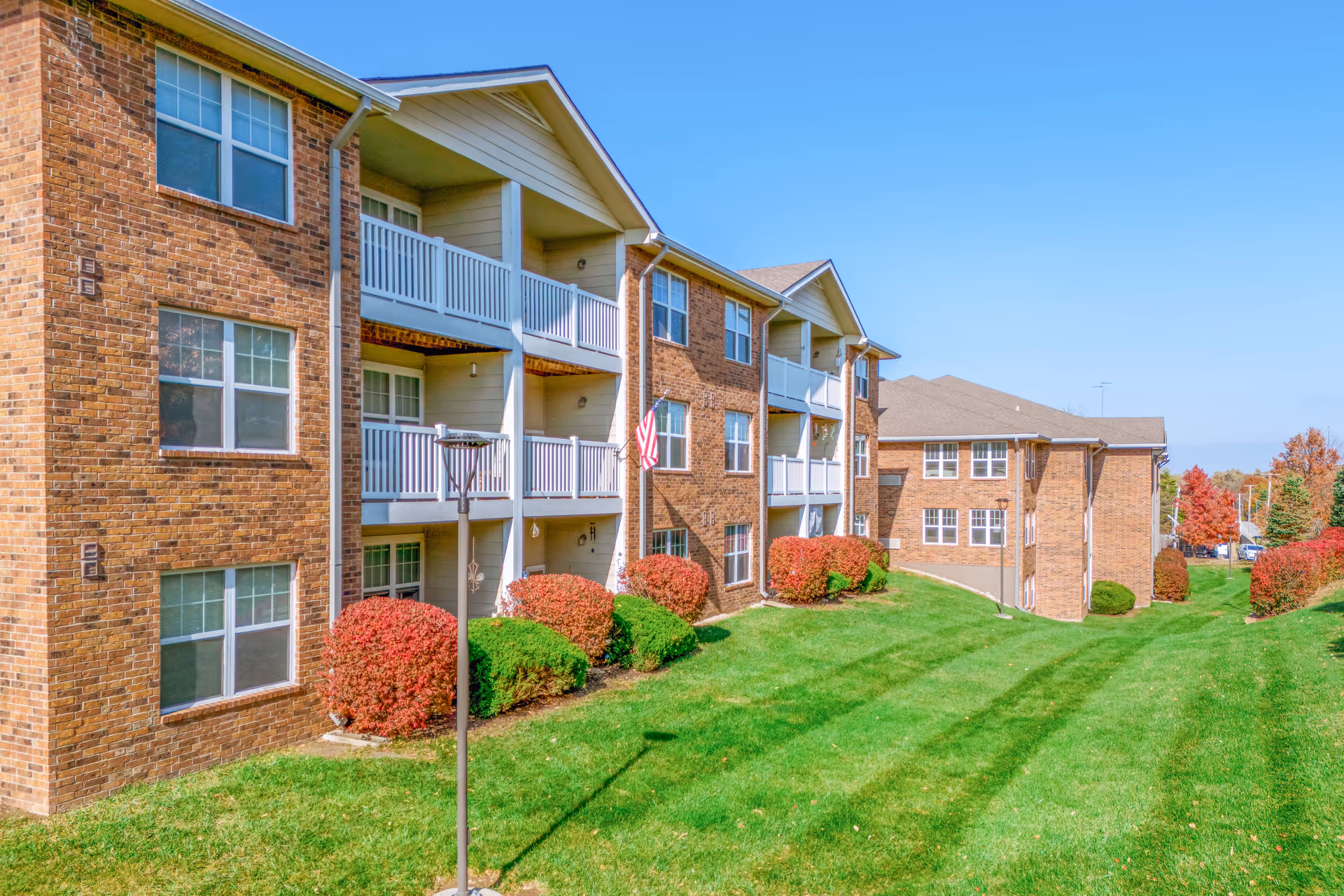 Brick senior living apartment building with balconies, green lawn, and trimmed shrubs.