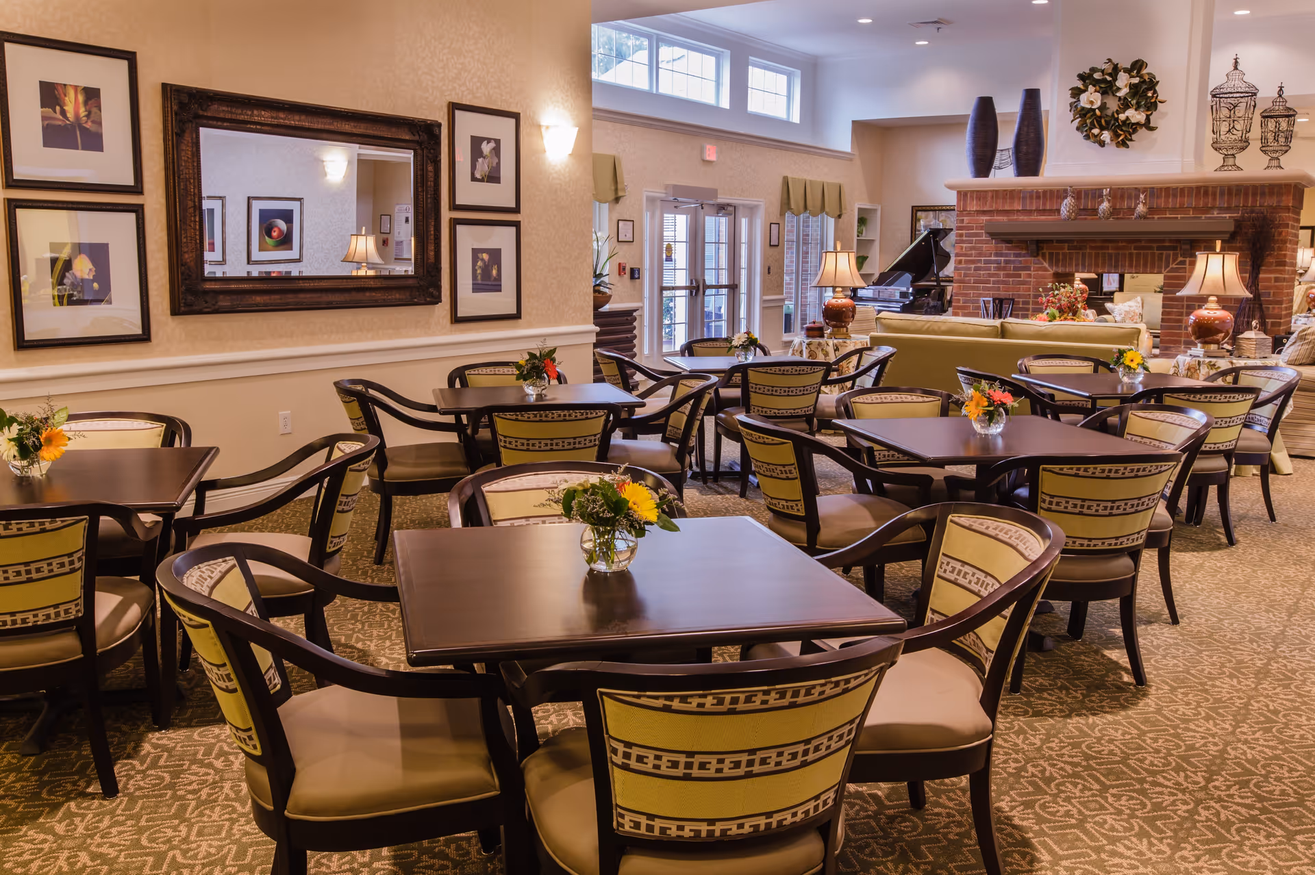 A cozy dining area in a senior living facility with multiple square wooden tables surrounded by cushioned chairs featuring patterned backs. Each table has a small vase with fresh flowers. The room has warm lighting, framed floral artwork on the walls, a large mirror, and a brick fireplace with decorative items on the mantel. Large windows and glass doors allow natural light to enter, and a grand piano is visible in the background.