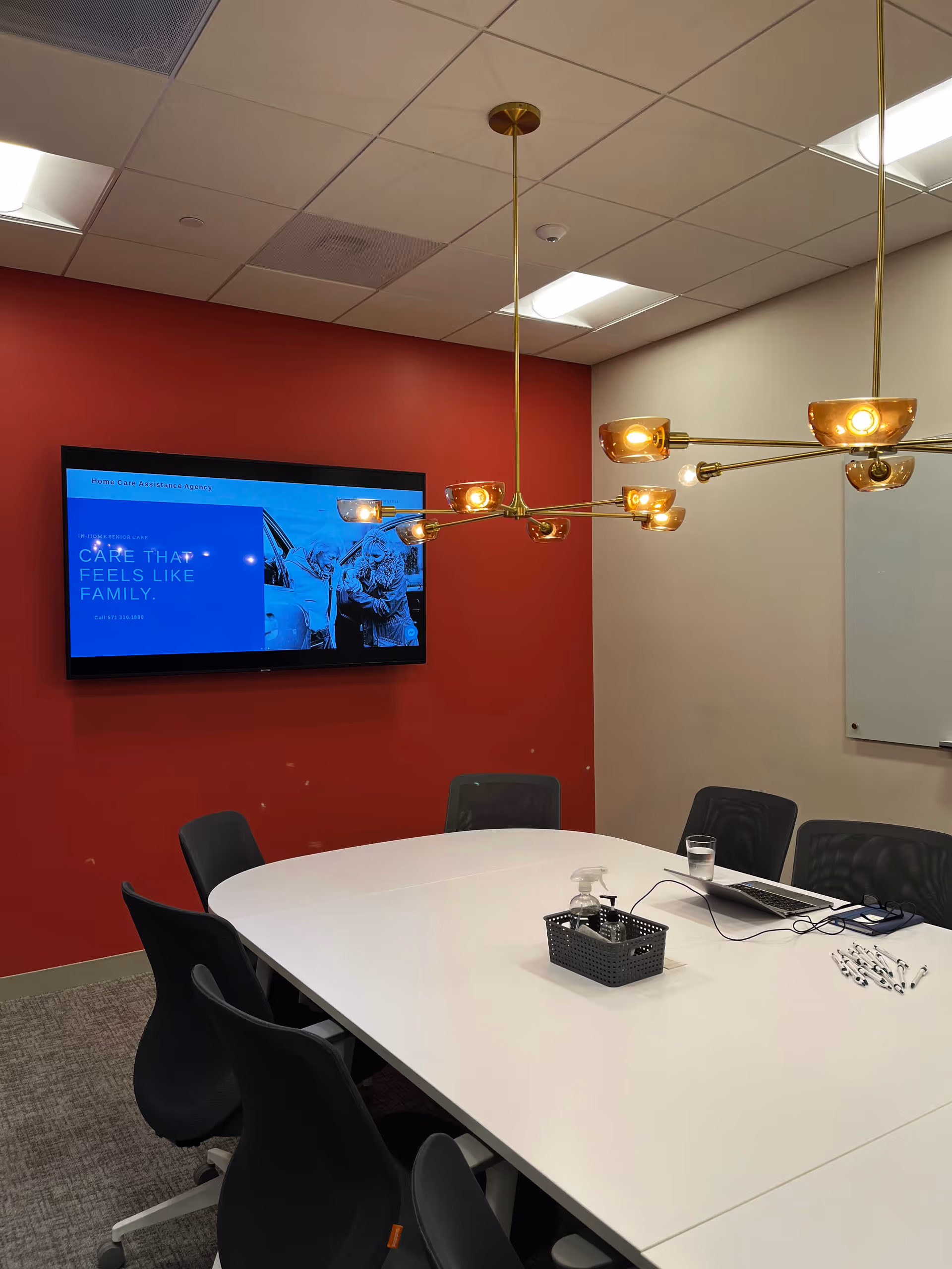 Conference room with white oval table, black chairs, red wall, and mounted screen.