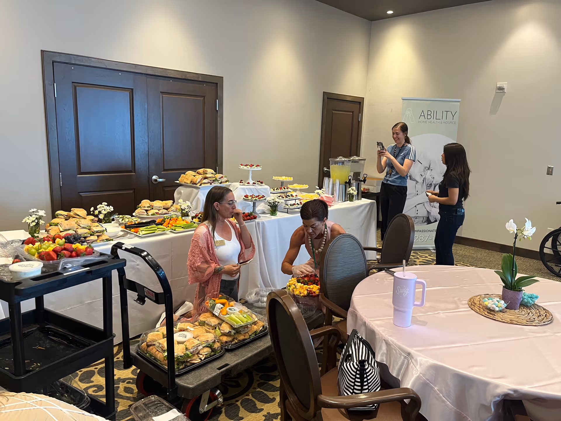 Staff and residents mingle near a buffet of sandwiches and desserts in a senior living facility