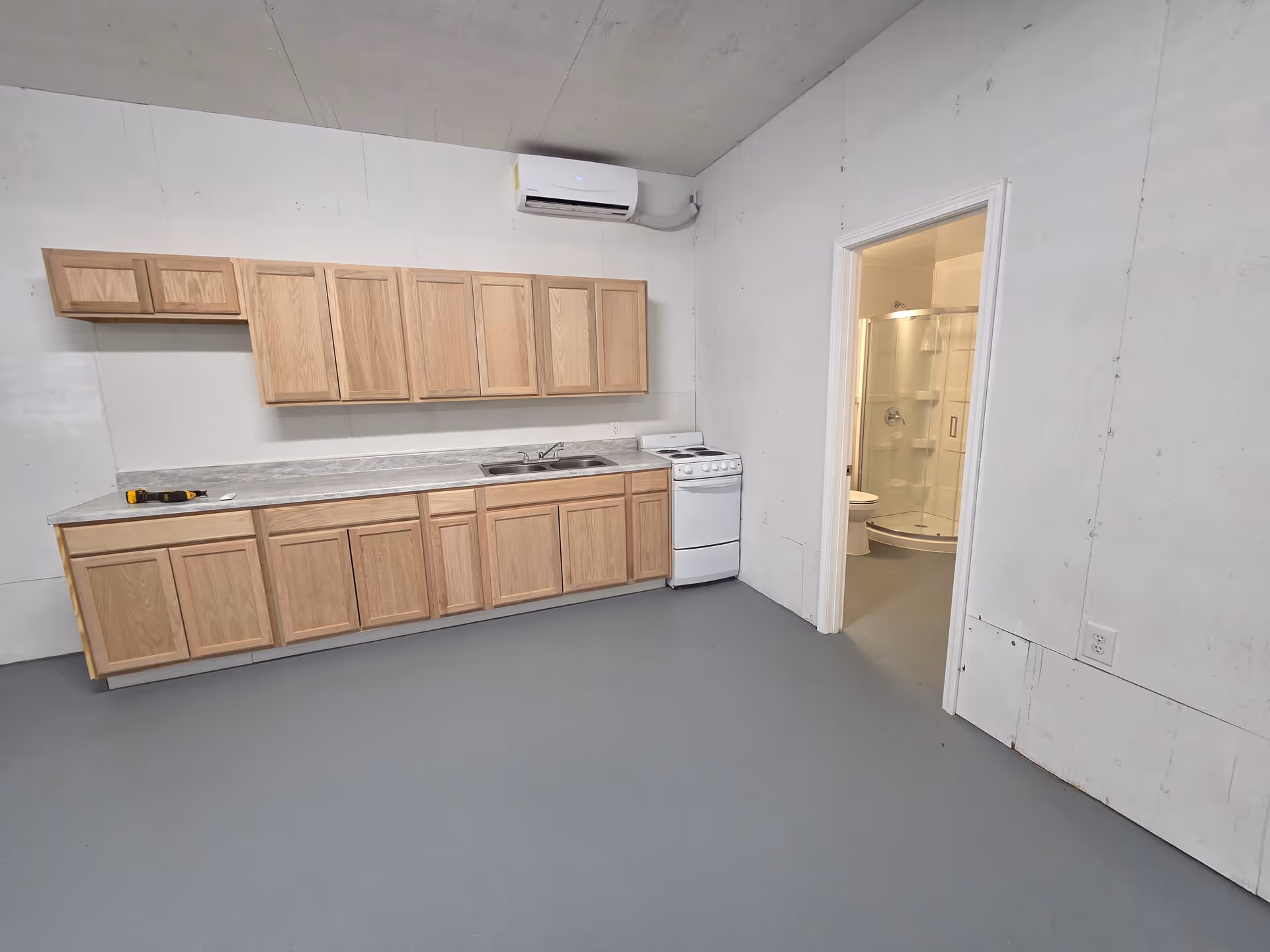 Empty utility kitchen with wooden cabinets, sink, stove, and bathroom doorway.