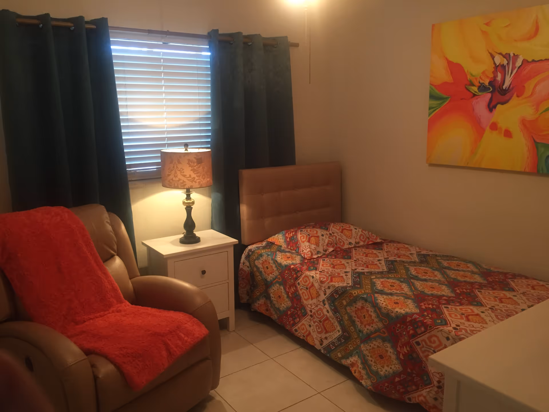 Cozy bedroom with a single bed in colorful patterned bedding, a recliner draped with a red throw, a nightstand and lamp by a curtained window, and bright wall art.