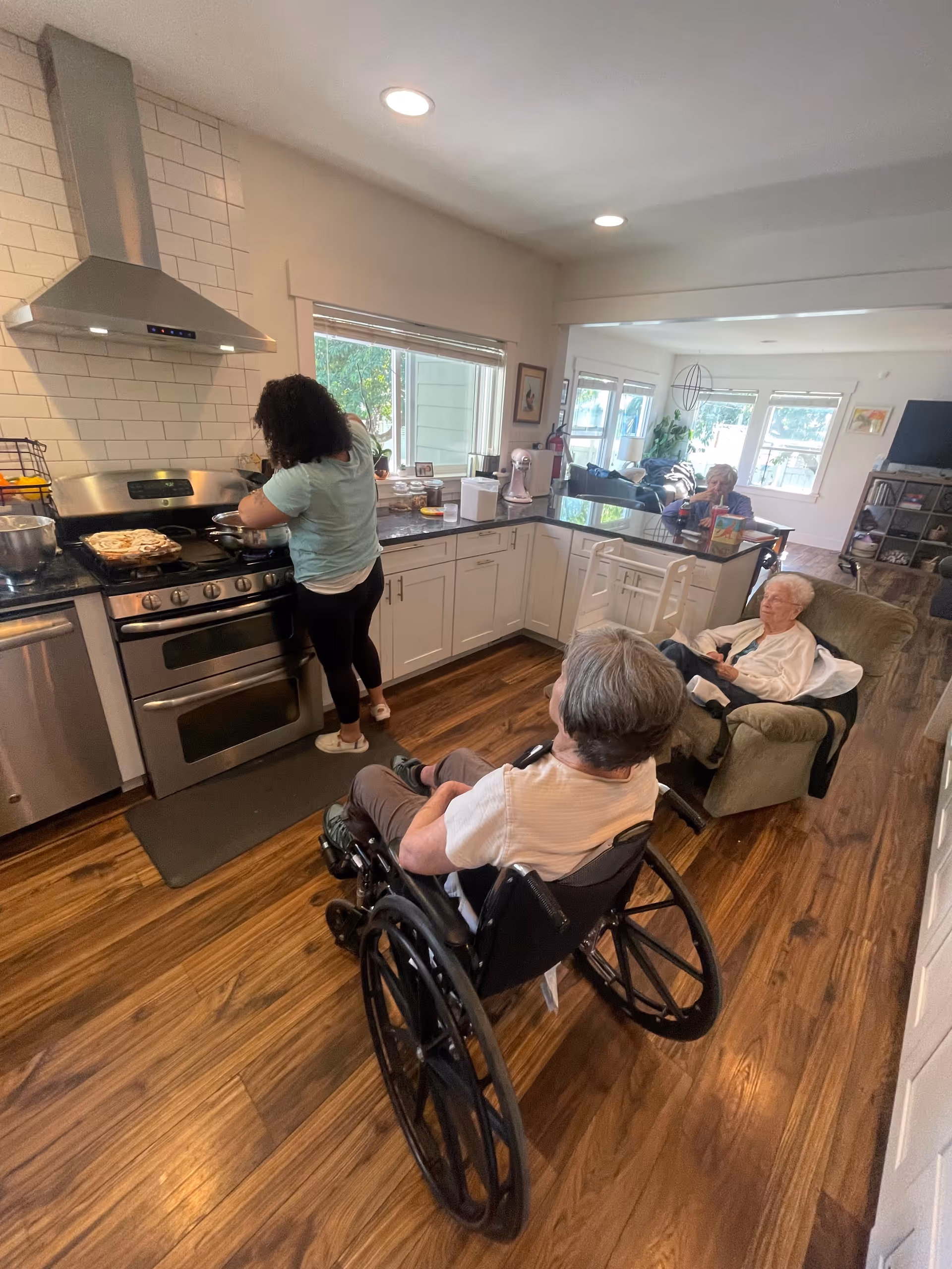 Caregiver cooks at stove in a bright kitchen while two elderly residents in wheelchairs sit nearby.