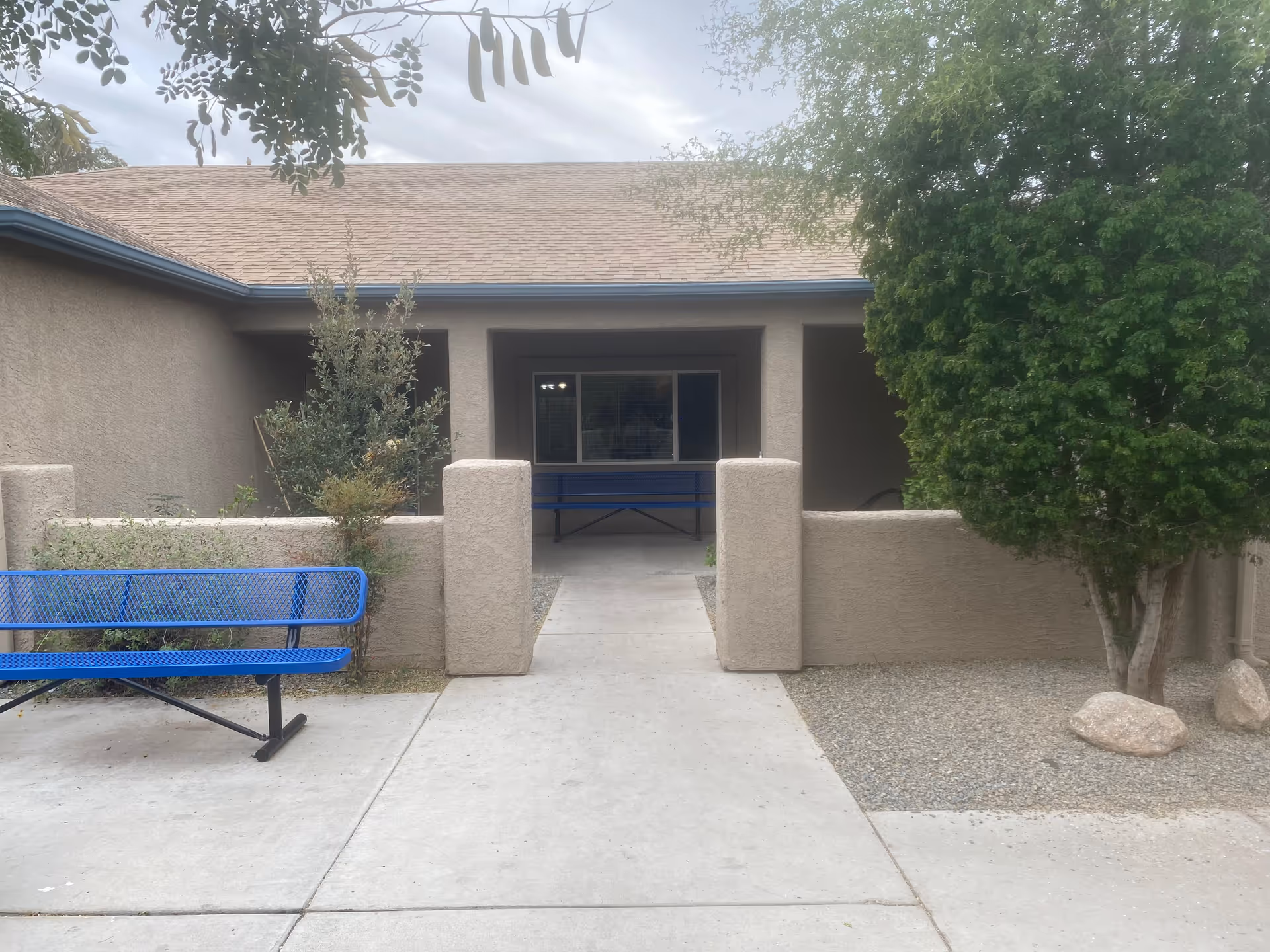 Entrance to a beige stucco building with a covered walkway, blue benches, shrubs, and a tree.