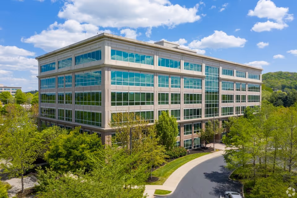 Modern glass-front office building surrounded by trees under a bright blue sky.