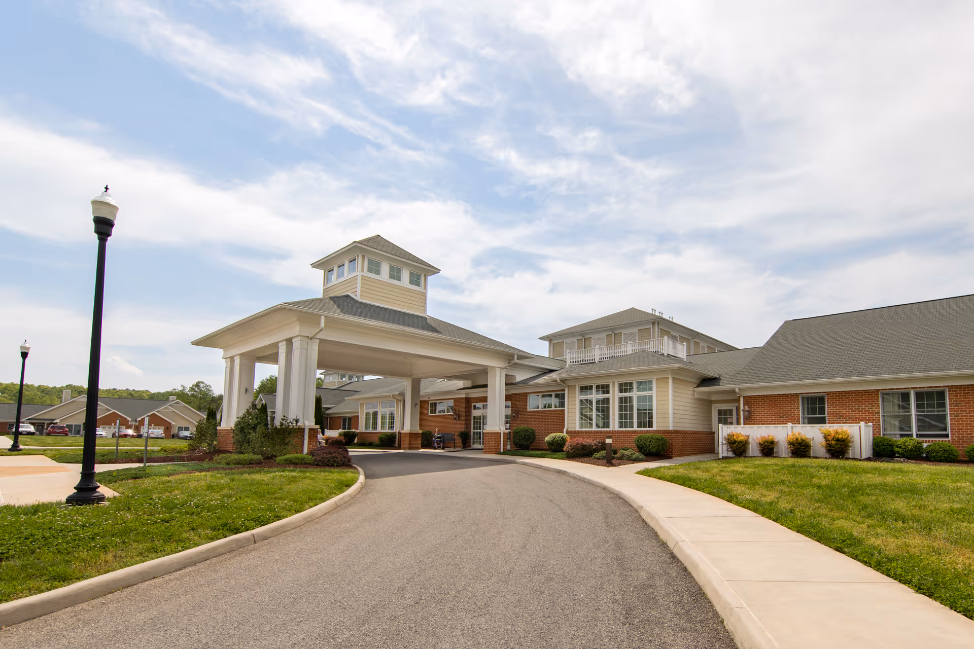 Exterior view of a senior living facility with a covered entrance driveway, brick and beige siding walls, multiple windows, and a well-maintained lawn under a partly cloudy sky.