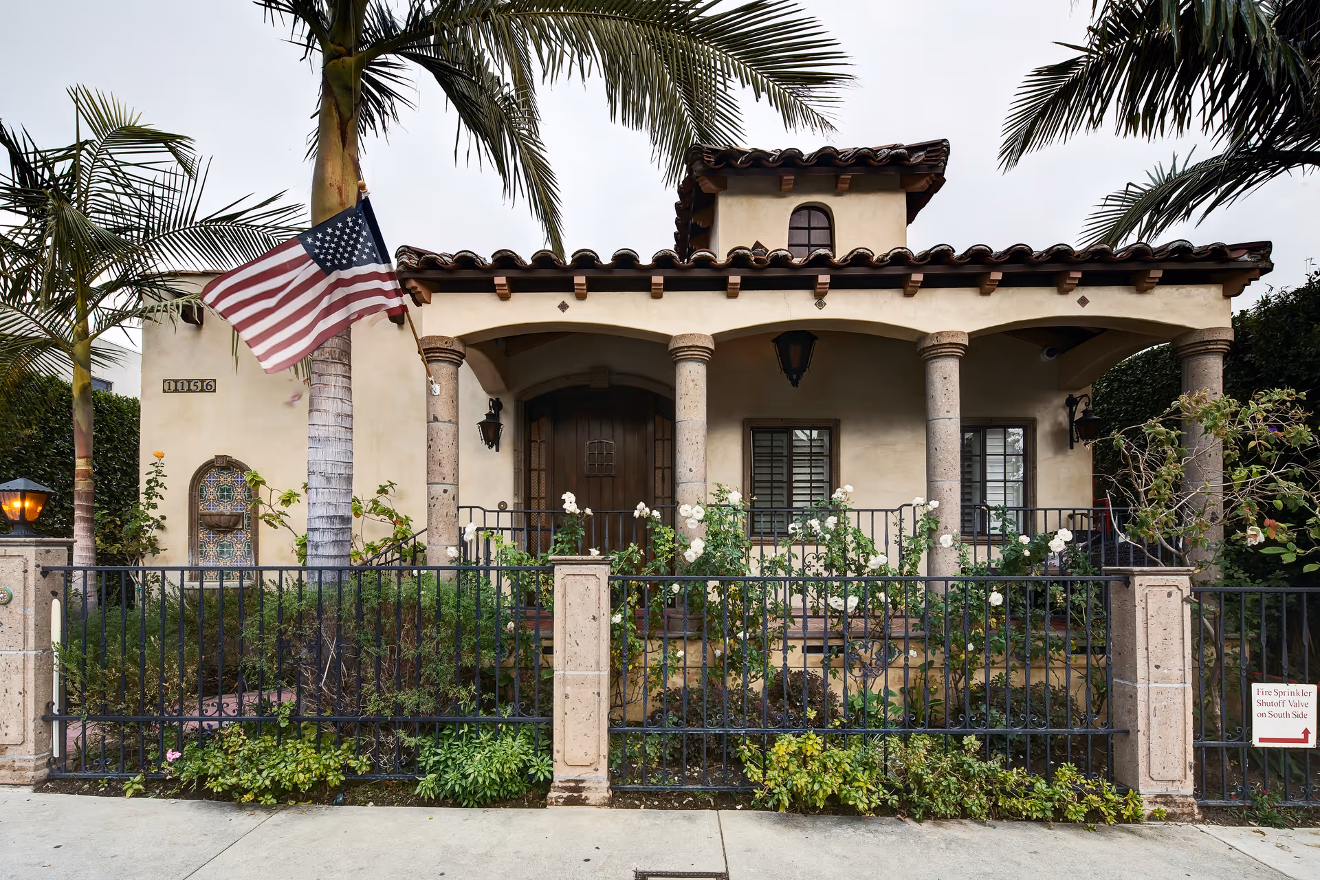 Front view of a Mediterranean-style facility with columns, gate, flowering garden, and flag.