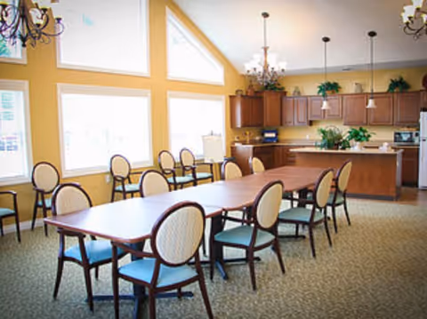 Bright communal dining room with a long table, multiple chairs, and a kitchen area in the background.