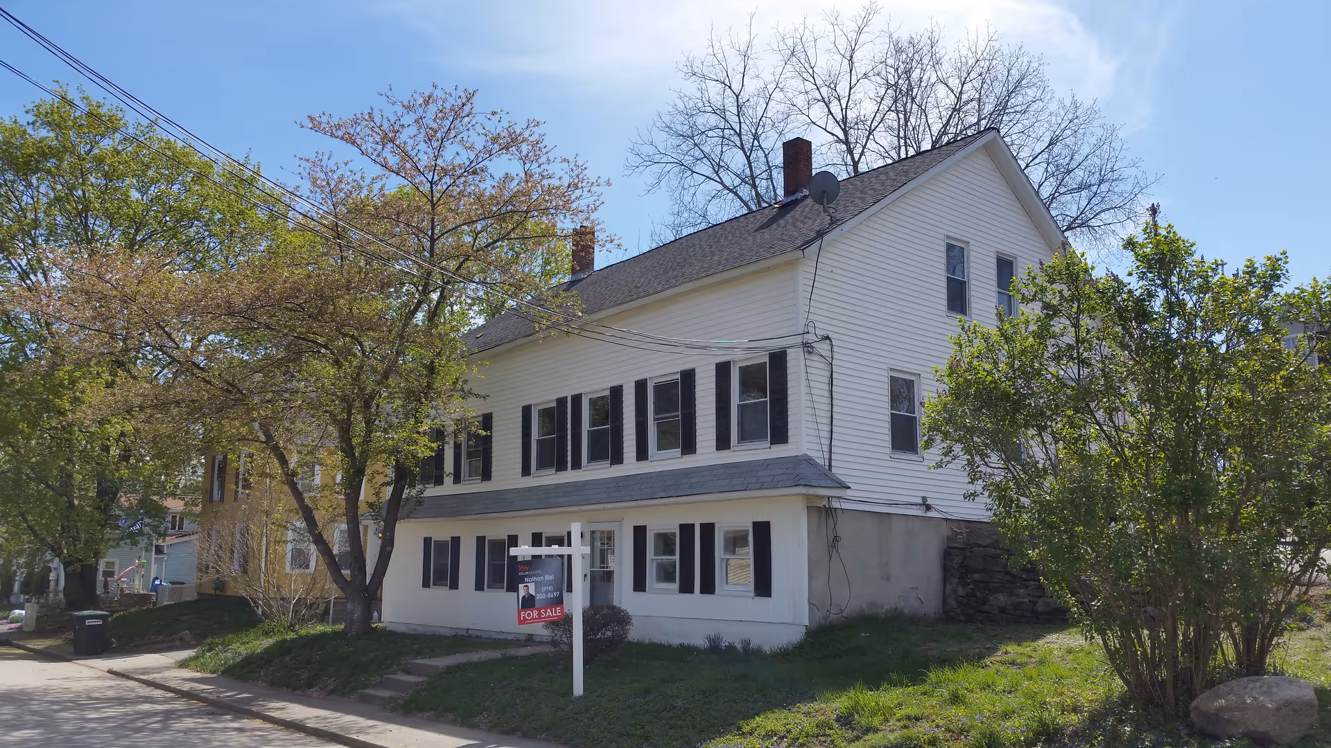 Two-story white building with black shutters and a For Sale sign in the front yard.