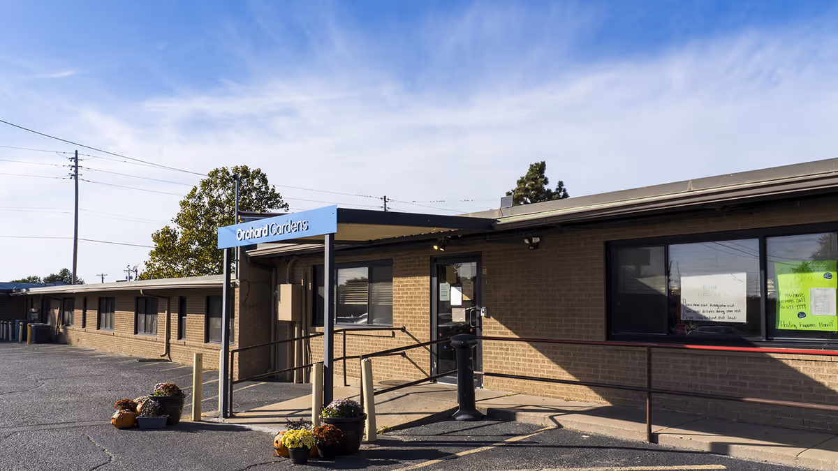 Single-story brick building entrance with a blue "Orchard Gardens" sign, accessibility ramp, potted plants, and a parking area under a clear sky.