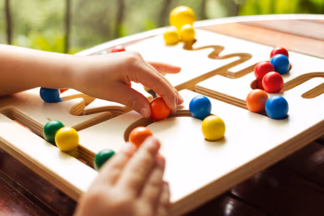 Hands guide colorful balls along a wooden tabletop labyrinth puzzle at a senior facility.