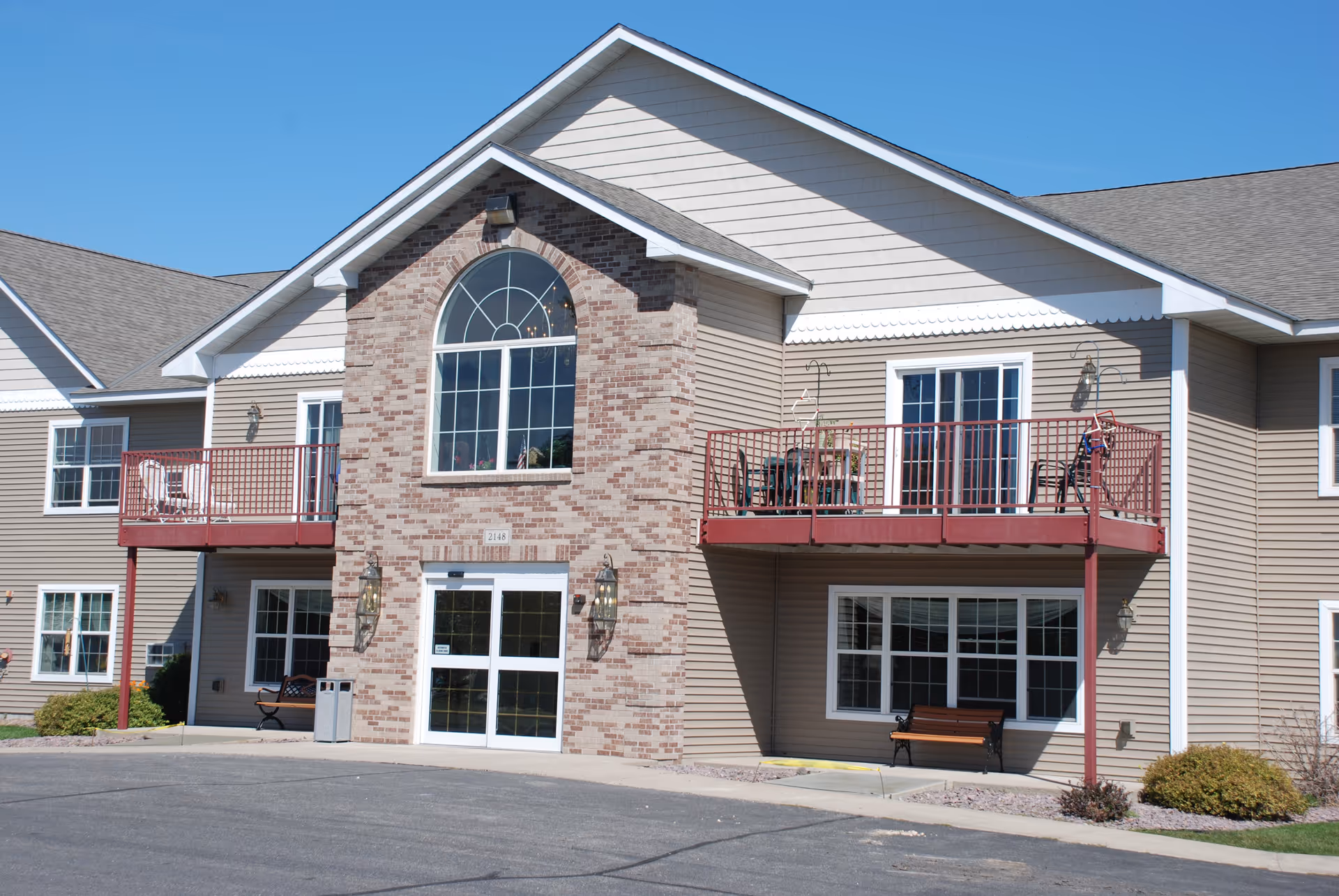 Exterior view of a two-story senior living facility building with beige siding and a central section of brick. The building features two red balconies with outdoor furniture, large windows, and a double glass entrance door. There are benches and some landscaping near the entrance under a clear blue sky.